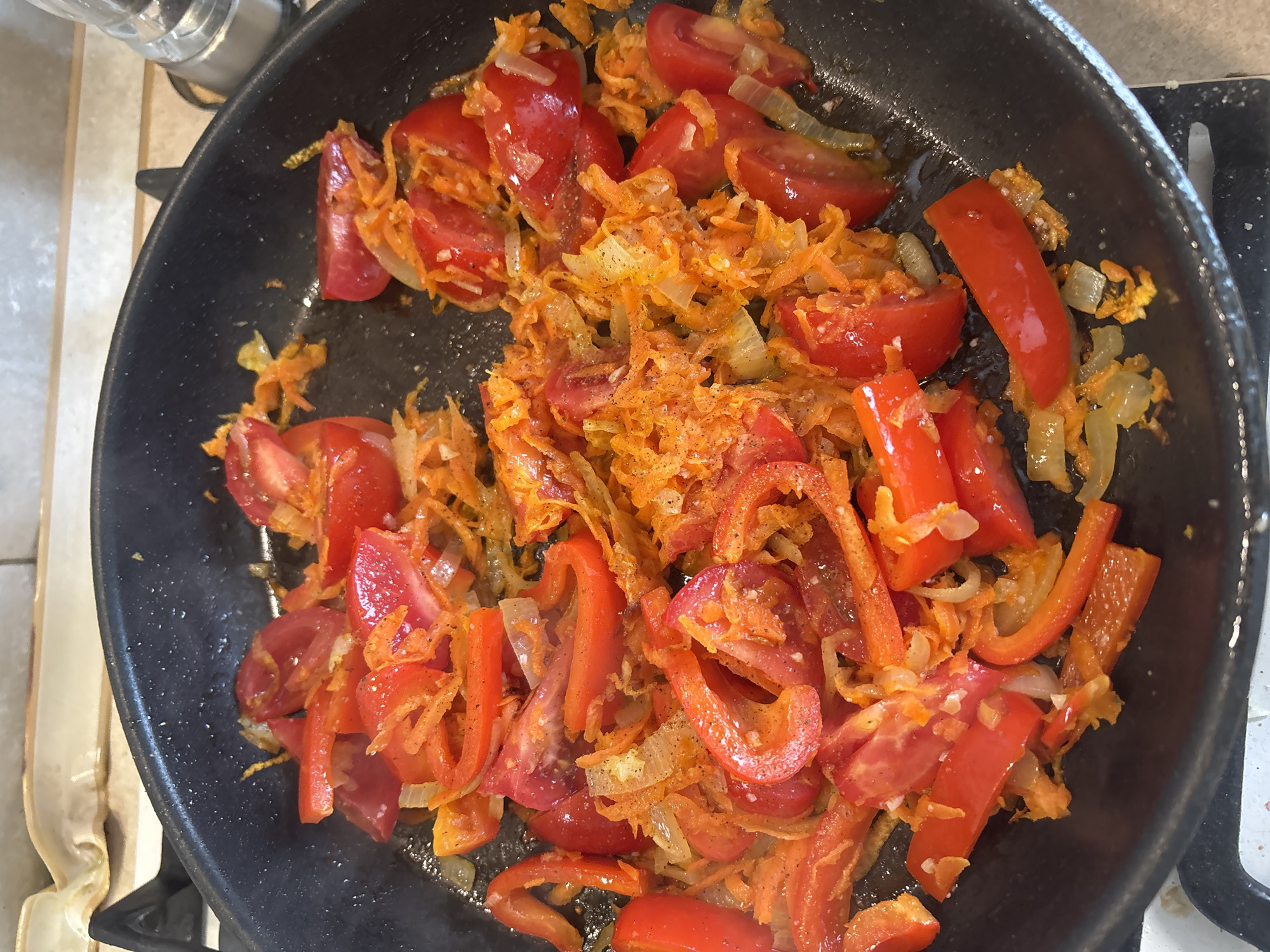 A close-up, overhead shot shows chopped tomatoes, shredded carrots, sliced onions, and red bell pepper pieces being sautéed in a black frying pan. The vegetables are in various stages of cooking, with some appearing softer and more translucent than others. The ingredients are seasoned with what looks like salt and pepper, and possibly some paprika, indicated by the reddish hue on some pieces. The pan is placed on a stovetop, with a sliver of a white countertop visible to the left and the edge of a black stovetop surface to the right. A clear glass container, possibly a salt or pepper shaker, is partially visible in the upper left corner. The lighting suggests an indoor setting, likely a kitchen, and the time of day is indeterminate. There is no visible text in the image. The overall scene depicts the preparation of a vegetable medley or a component of a larger dish.