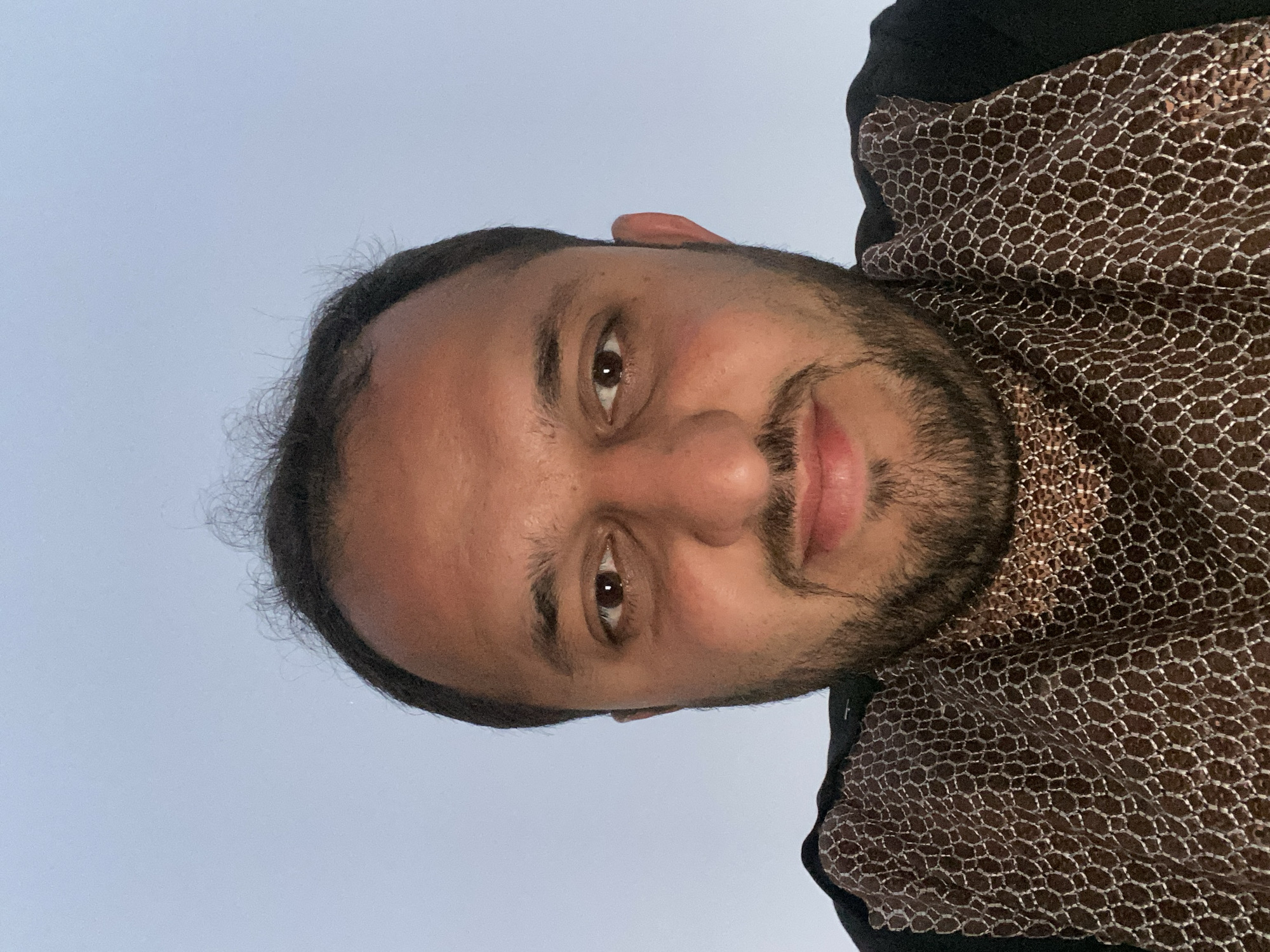 A man with dark hair and a beard is centered in the frame, looking directly at the camera. He has brown eyes, dark eyebrows, and slightly parted lips. He is wearing a brown and silver patterned shirt over a black garment. The background is a light blue, likely the sky. There is no indication of the location being Kandahar, Afghanistan.