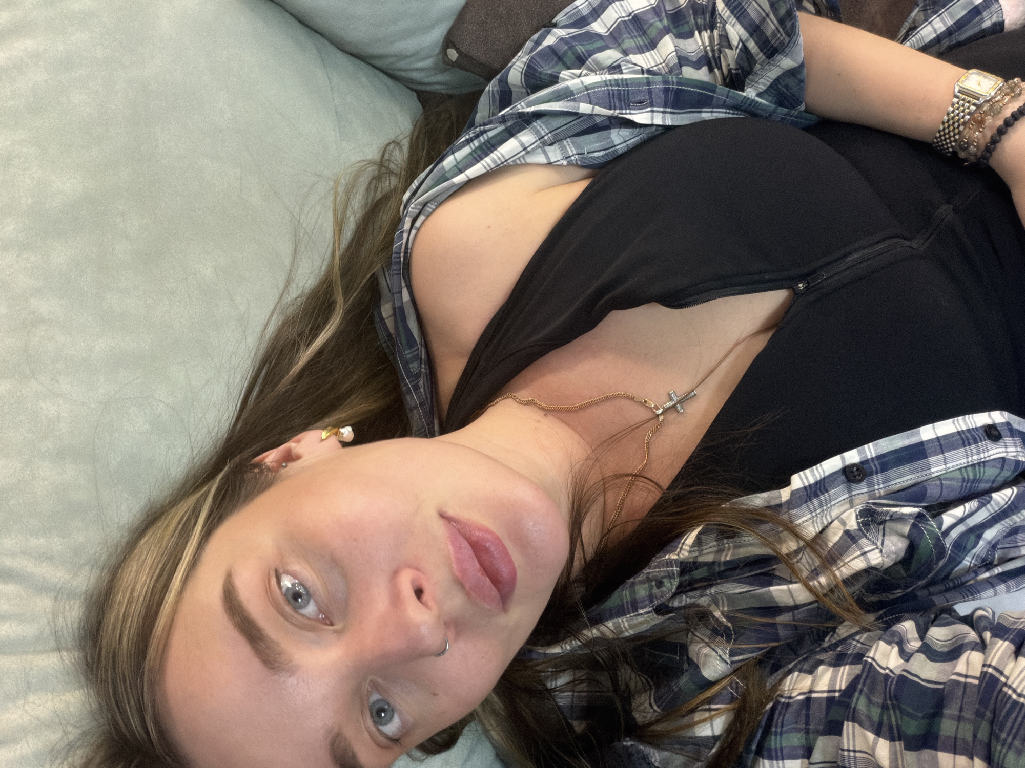 A young woman is captured in a close-up, angled shot, lying on what appears to be a light green/teal sofa or bed. Her long, light brown hair with blonde highlights is spread around her head. She has light blue eyes and a neutral expression, with slightly parted, plump lips and a nose ring. She wears a black top, partially covered by an unbuttoned blue, green, and white plaid shirt. Her accessories include a delicate gold chain necklace with a small, sparkling cross pendant, a gold wristwatch on her right wrist, and multiple beaded bracelets. She also has a pearl stud earring in her left ear and another gold-colored earring. The background shows the textured fabric of the furniture and a darker cushion near her head. The woman is looking directly at the camera, suggesting a selfie or posed photograph. The setting is indoors in Chaikovsky, Russia.
