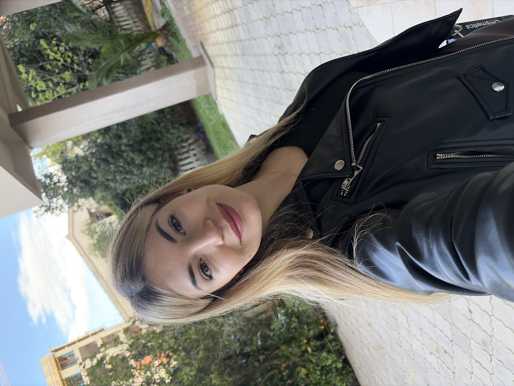 The image shows a woman with blonde hair, wearing a black leather jacket. She is outdoors, likely in the city of Budva, Montenegro. Buildings and trees can be seen in the background. The ground is covered in paving stones.