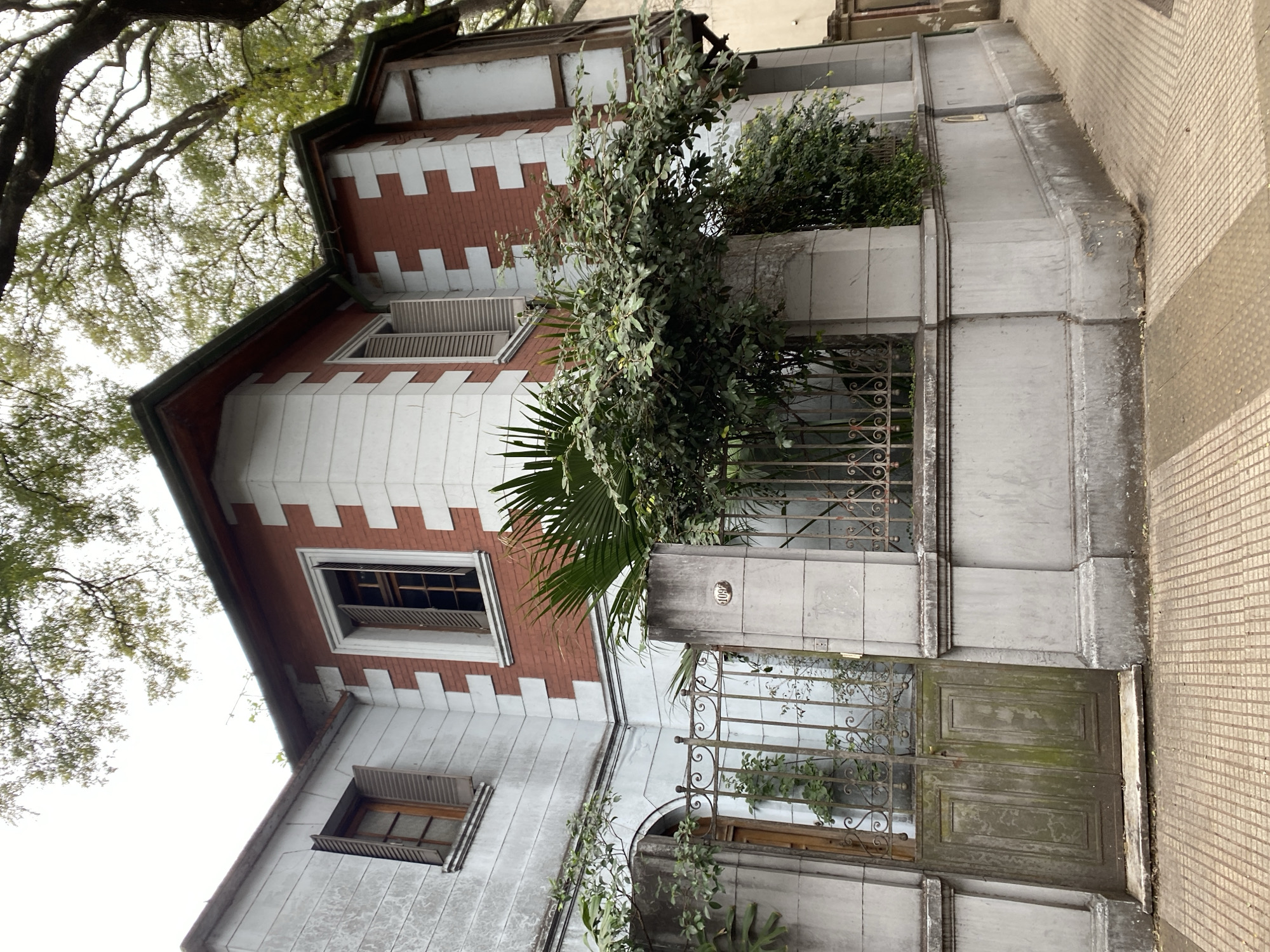 The image shows a red and white brick house with a gray stone facade on a street in Buenos Aires, Argentina. The house has several windows with wooden shutters, some of which are open. There is a wrought-iron gate in front of the house, and a walkway made of interlocking square tiles leads to the gate. Trees with green leaves are visible in the background, and the sky is overcast. The house appears to be old and somewhat dilapidated, with peeling paint and moss growing on the gate. The number "109" is visible on a plaque near the gate.