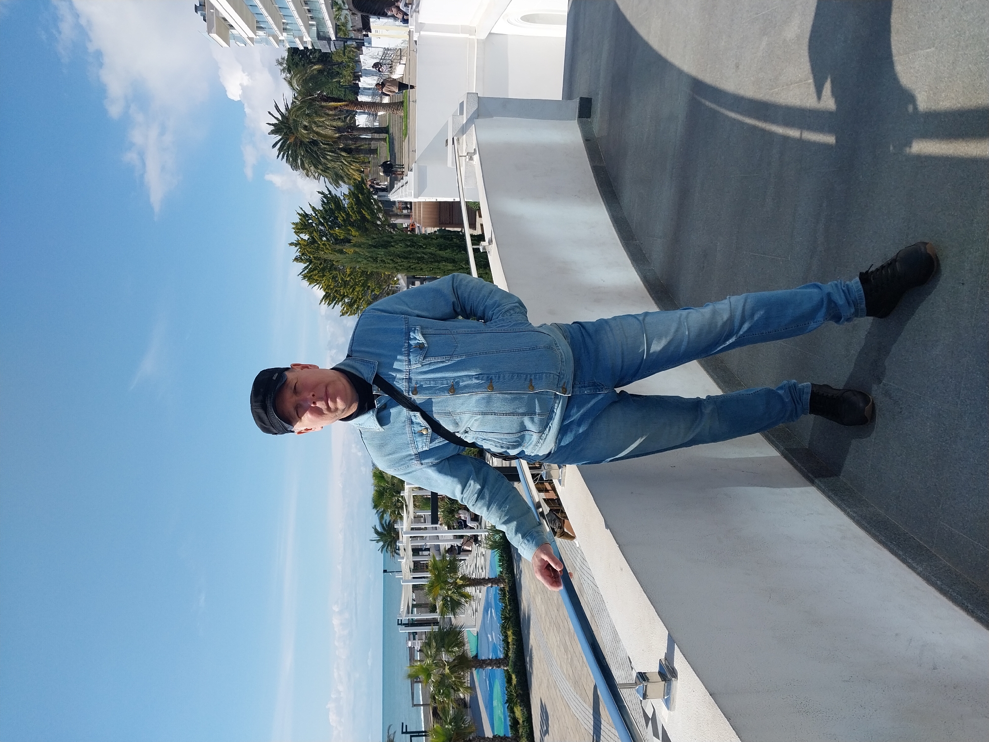 A man wearing a denim jacket, jeans, black shoes, and a black cap is standing on a white concrete platform with a blue railing. The platform overlooks a pool area with palm trees and a building in the background. To the left of the man is a clear blue sky with a few clouds. A shadow cast by his legs is visible on the grey concrete surface to his right. The scene is likely in Sochi, Russia, judging by the architecture and landscape.