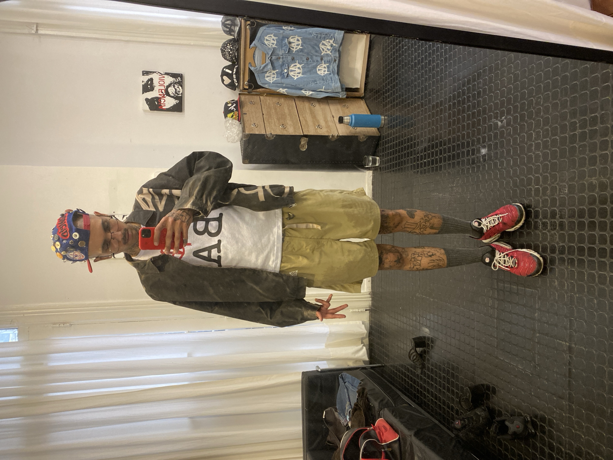 This mirror selfie, taken in Buenos Aires, Argentina, features a person dressed in a casual outfit, posing in what appears to be a bedroom or dressing area. The individual, who has visible tattoos on their arms and legs, is wearing glasses and a distinctive blue hat covered with various buttons. Their attire includes a dark jacket, a white t-shirt with the letters "BA" printed on it, khaki shorts, dark socks, and red and white sneakers. They are holding a red smartphone, presumably used to capture the image.
The background of the shot reveals several details of the room. To the right of the person, an open, vintage-style wooden and black trunk displays a light blue denim jacket adorned with white anarchist symbols, and several black and white hats. A blue water bottle is placed on the trunk. On the white wall behind, a framed piece of art depicts a stylized face and the reversed text "MICHAEL JACKSON." The floor is covered with a textured black mat. To the person's left, white window blinds are visible, and in the foreground, a black bench or sofa holds a red and black bag. The room is brightly lit, suggesting the photo was taken during the day.