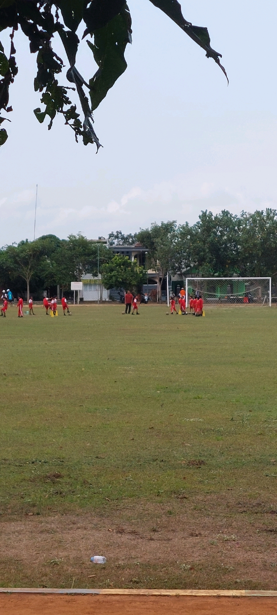 A group of children in red uniforms are on a grassy field, appearing to practice soccer. They are scattered across the field, some near a goalpost on the right side of the image, while others are in smaller groups or spread out individually. There are also a few adults present, also in red, seemingly supervising or coaching. The field is wide and mostly green, with some patchy dry areas. In the background, there are trees and buildings, suggesting an urban or suburban setting. The sky is overcast. A discarded plastic water bottle is visible on the grass in the foreground.