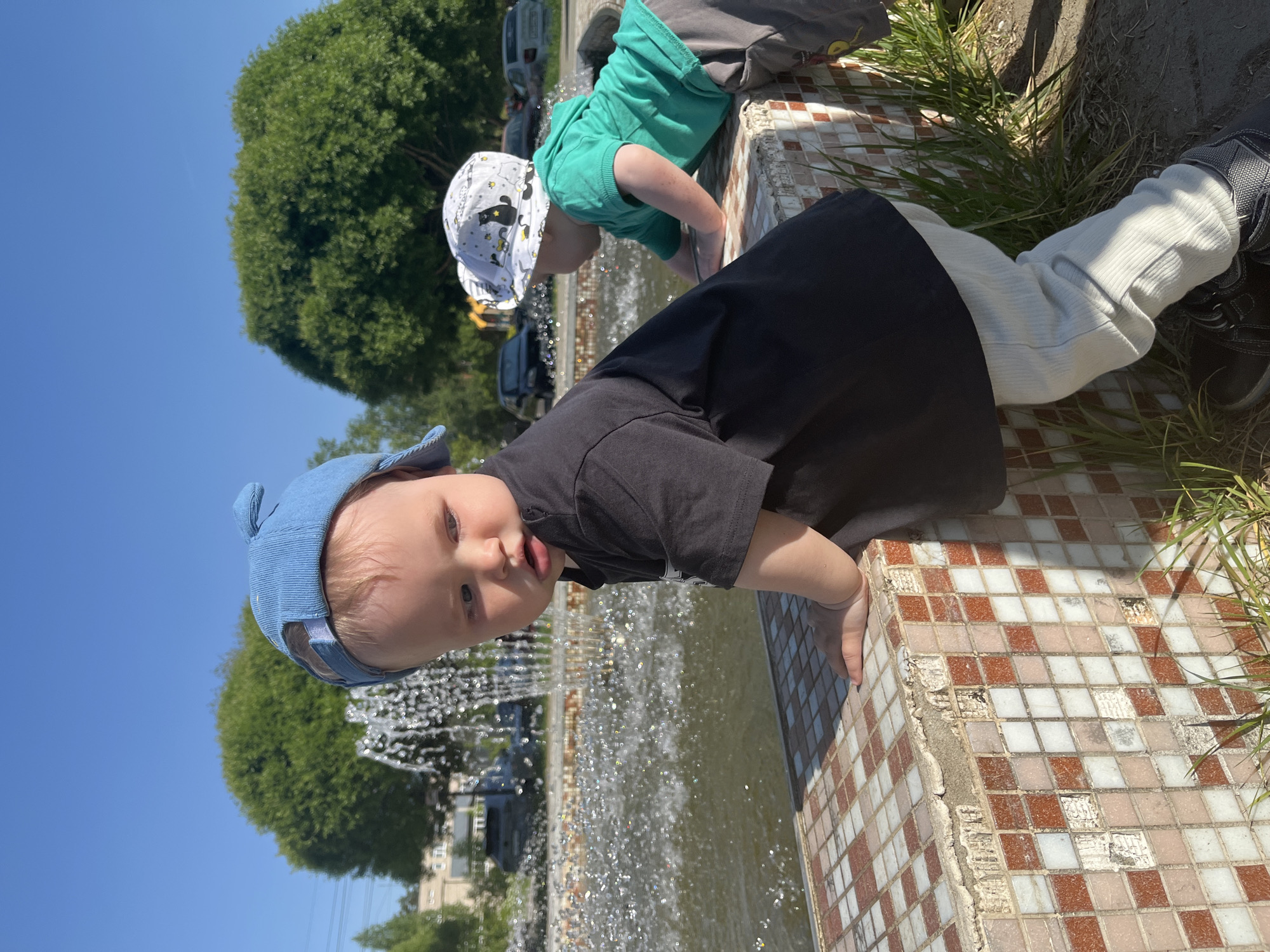 Two young children are playing at the edge of a fountain. One child wears a blue cap and a black shirt. The other child is in a green shirt and a white hat. The fountain is tiled with small, square tiles. Trees and buildings are visible in the background. The photograph may have been taken in the city of Chaikovsky, Russia.