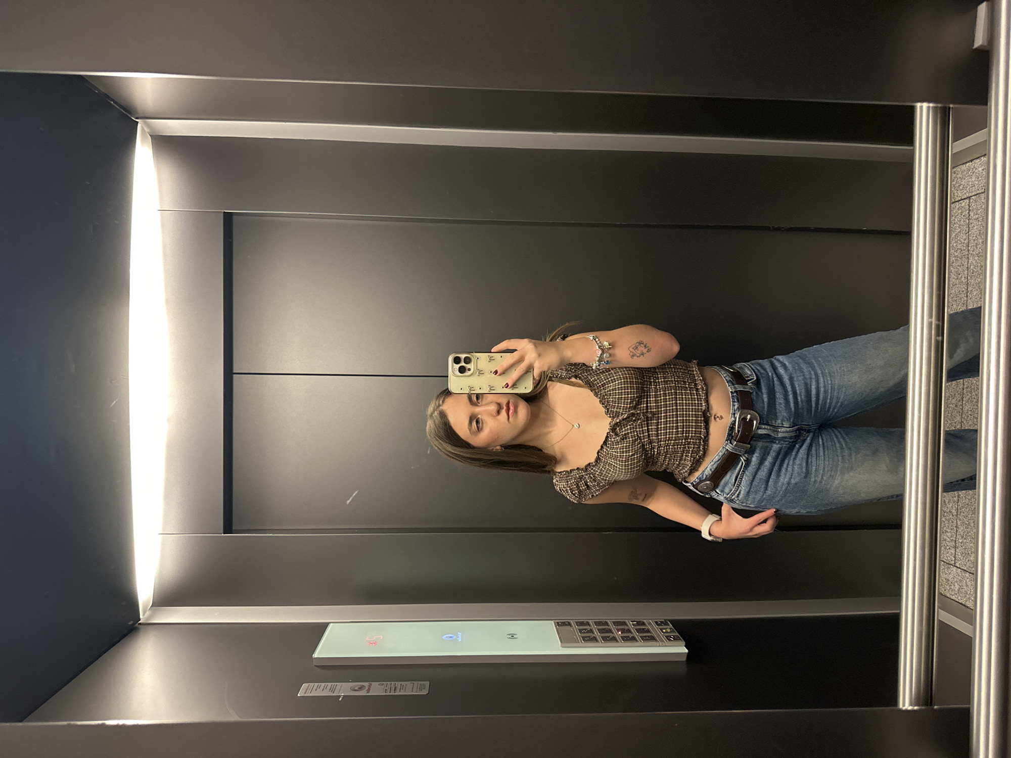 A young woman is depicted taking a mirror selfie inside a modern elevator. The image is rotated 90 degrees counter-clockwise. She has long, dark hair with lighter highlights and is wearing a brown and white plaid, ruffled crop top, blue jeans, and a dark brown belt with a prominent buckle. Her left hand holds a light-colored smartphone with a decorative case, capturing her reflection as she looks directly at the camera. She has multiple bracelets on her left wrist, a white smartwatch on her right wrist, and a visible tattoo on her left forearm. A small belly button piercing is also apparent.
The elevator interior features dark gray walls and a bright, vertical light strip on the left side. A shiny metal handrail runs along the right wall. At the bottom of the reflection, a sleek elevator control panel is visible, displaying illuminated buttons and a digital screen showing "1" and "H." The floor has a speckled gray and white pattern. The scene takes place indoors, likely in a building in Huixquilucan de Degollado, Mexico. The lighting indicates an indoor setting, not necessarily specific to a time of day.