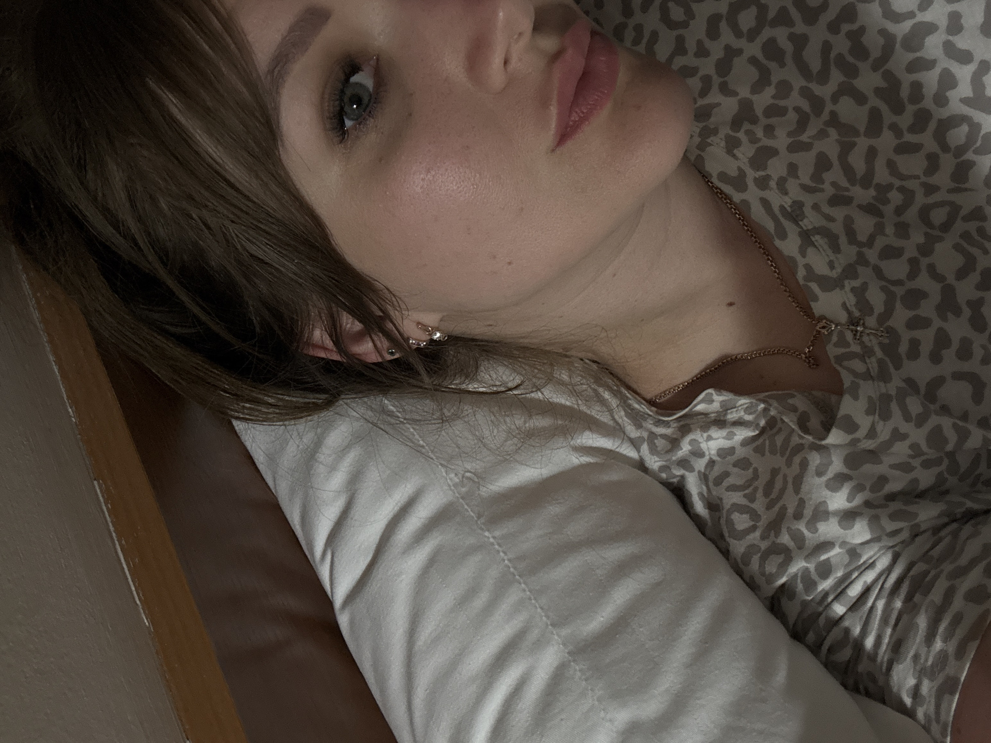 A woman with light skin, brown hair, and blue eyes is lying on a white pillow. She is wearing a gray and white animal print shirt and a gold necklace with a cross pendant. A wooden object is visible on the left side of the image. The location is not discernible in this image.