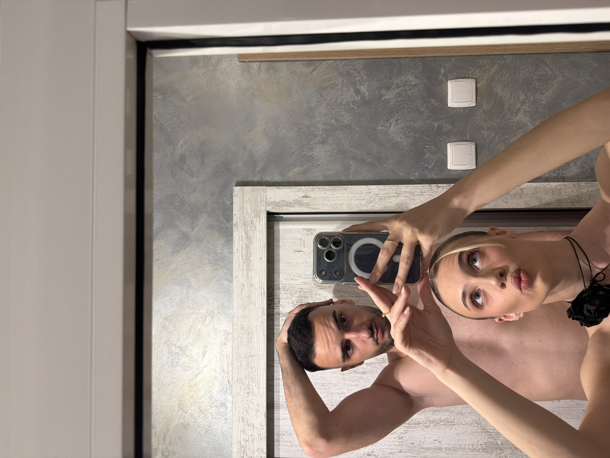 A horizontally-oriented, medium shot depicts two partially nude individuals taking a selfie in front of a reflective surface. The lighting is diffused.
The male figure, with dark hair and facial stubble, holds his hand on his head. The female figure, with blonde hair pulled back, puckers her lips. A black floral ornament is visible around her neck.
A smartphone with a clear case is held in front of them. The background consists of a gray, textured wall, two light switches, and a white door frame. The location and city are not deducible from the image.