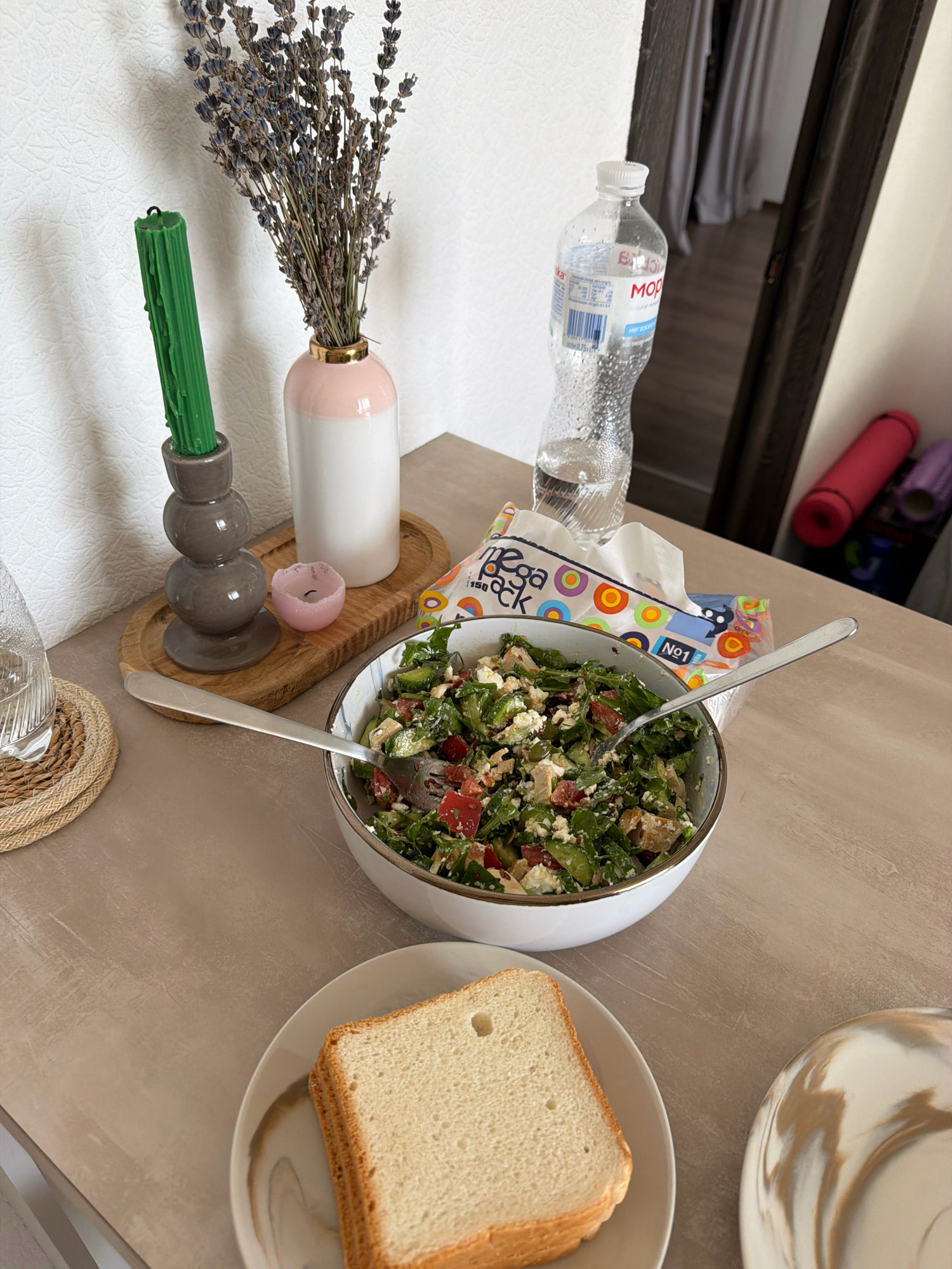 The image depicts a close-up, top-down view of a dining table set for a meal. The table is a light brown, textured surface. In the center of the frame, a white bowl contains a colorful salad with greens, tomatoes, and cheese. A fork rests inside the bowl, indicating the meal is in progress. To the left of the salad, a slice of white bread sits on a patterned plate.
Further left, a woven coaster holds a glass of water. Behind the coaster, a wooden tray holds a green candle in a grey candlestick, a small pink candle, and a vase containing dried lavender. A bottle of water is visible in the background, to the right of the lavender. The background includes a doorway with curtains and a shelf with rolled-up mats, possibly yoga mats.
The scene is set indoors, likely in a residential setting. The lighting suggests it is daytime, with natural light entering from the side. There are no people visible in the image, and the focus is on the food and decorative items on the table. The overall impression is one of a simple, healthy meal. The city of Kyiv, Ukraine, is not explicitly identified in the image.