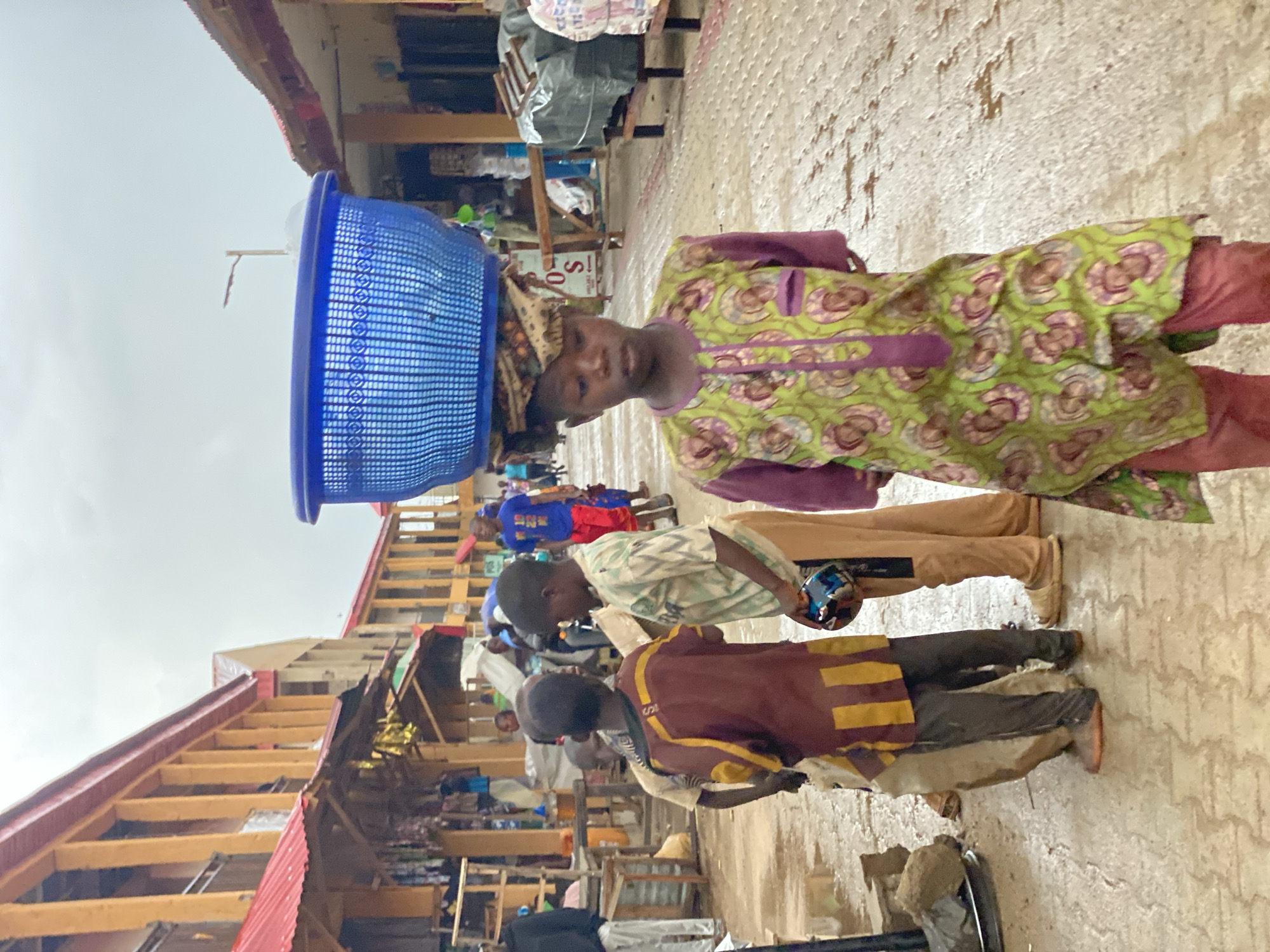 The image depicts a street scene in Kafanchan B, Nigeria. A person wearing a green and purple patterned top and red pants balances a blue basket on their head. Several other individuals are visible behind them, including two children. The surrounding environment includes buildings with a red roof and a partially covered market area. The ground appears wet.