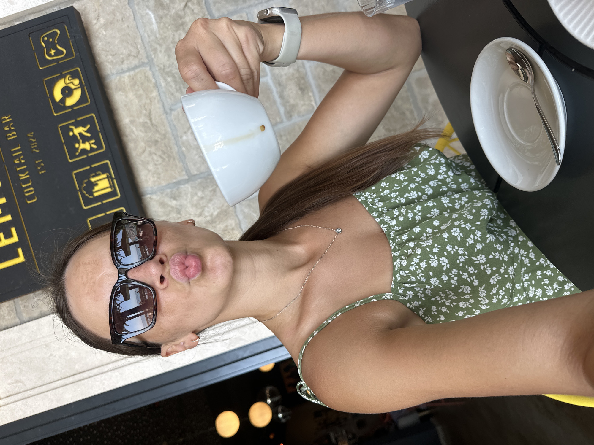 A young woman with brown hair, wearing sunglasses and a green floral dress, sits at a black table in an outdoor cafe. She is holding a white coffee cup with a small amount of coffee residue visible on the inside. Her right hand, adorned with a white smartwatch, is positioned near the cup. In front of her on the table is a white saucer with a spoon. The background includes a textured stone wall and a dark awning with yellow lettering that reads "COCKTAIL BAR EST 2024" and features various yellow icons. The scene appears to be in the city of Bečići, Montenegro.