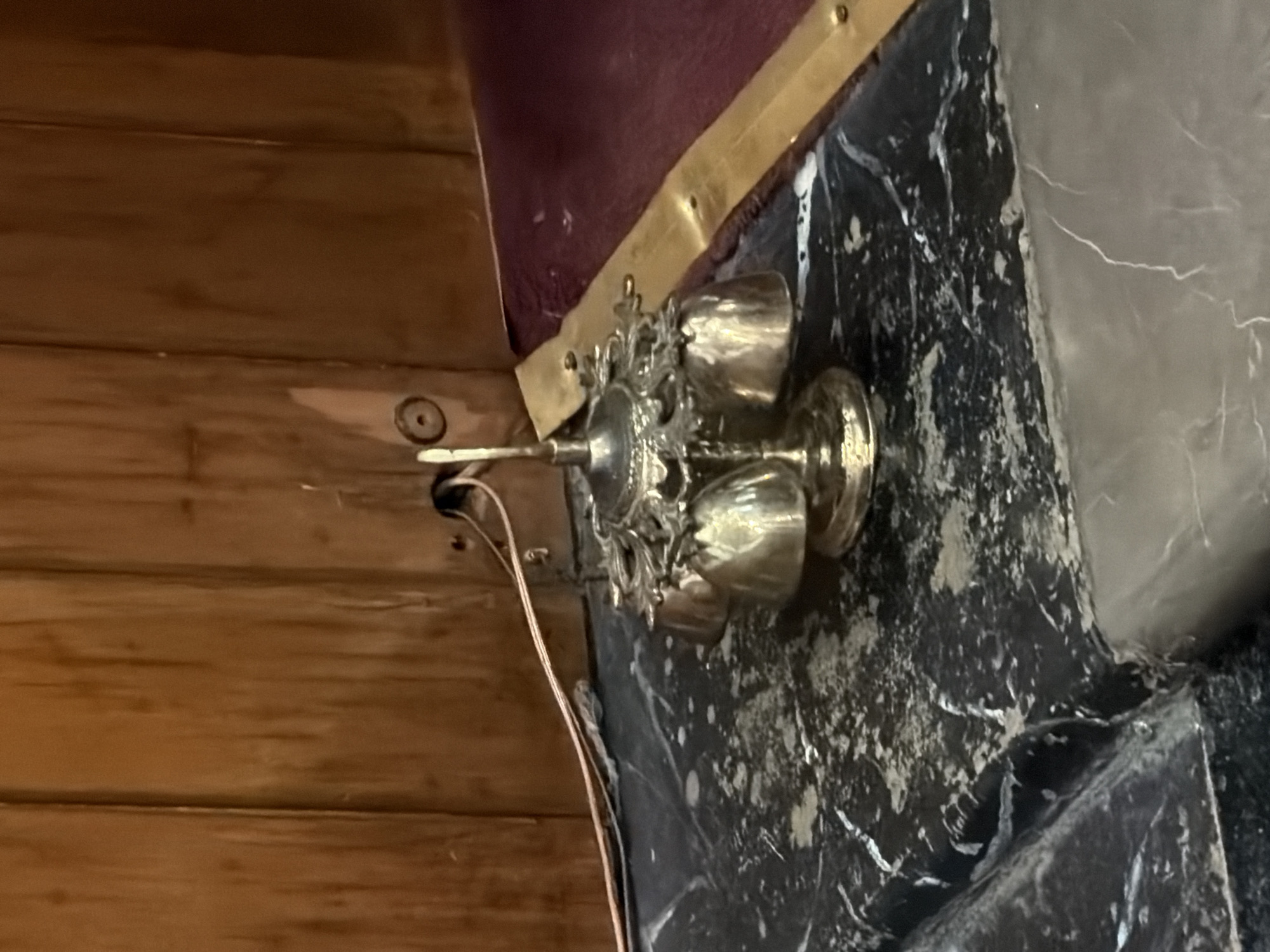 A close-up, somewhat angled shot shows a silver candelabra with several cup-shaped candle holders affixed to a wall with a dark, marbled surface. The candelabra is mounted on the corner where this wall meets a wooden wall. Visible wires emerge from the wall near the candelabra. A golden trim runs along the edge above the marbled wall. The location is Puebla, Mexico.