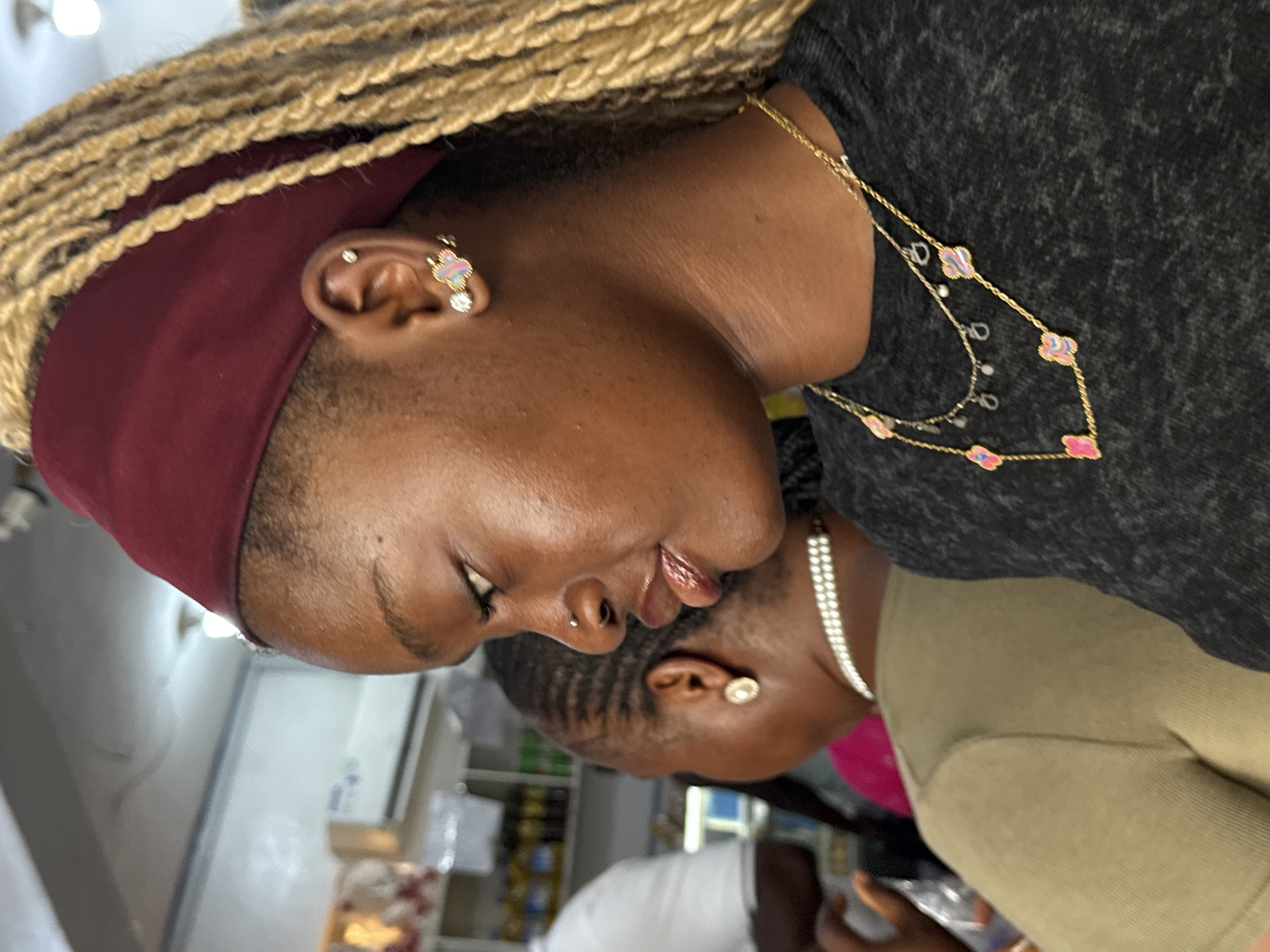 The image shows two Black women in an interior setting. The woman in the foreground has blonde braided hair with a burgundy headband. She is wearing a dark gray t-shirt and a gold necklace with multi-colored clover charms. She has multiple ear piercings with earrings and a nose piercing. The second woman has dark braided hair and is wearing a pearl necklace and olive green shirt. The background shows shelves with various items suggesting a store or shop. The location is Ado, Nigeria.