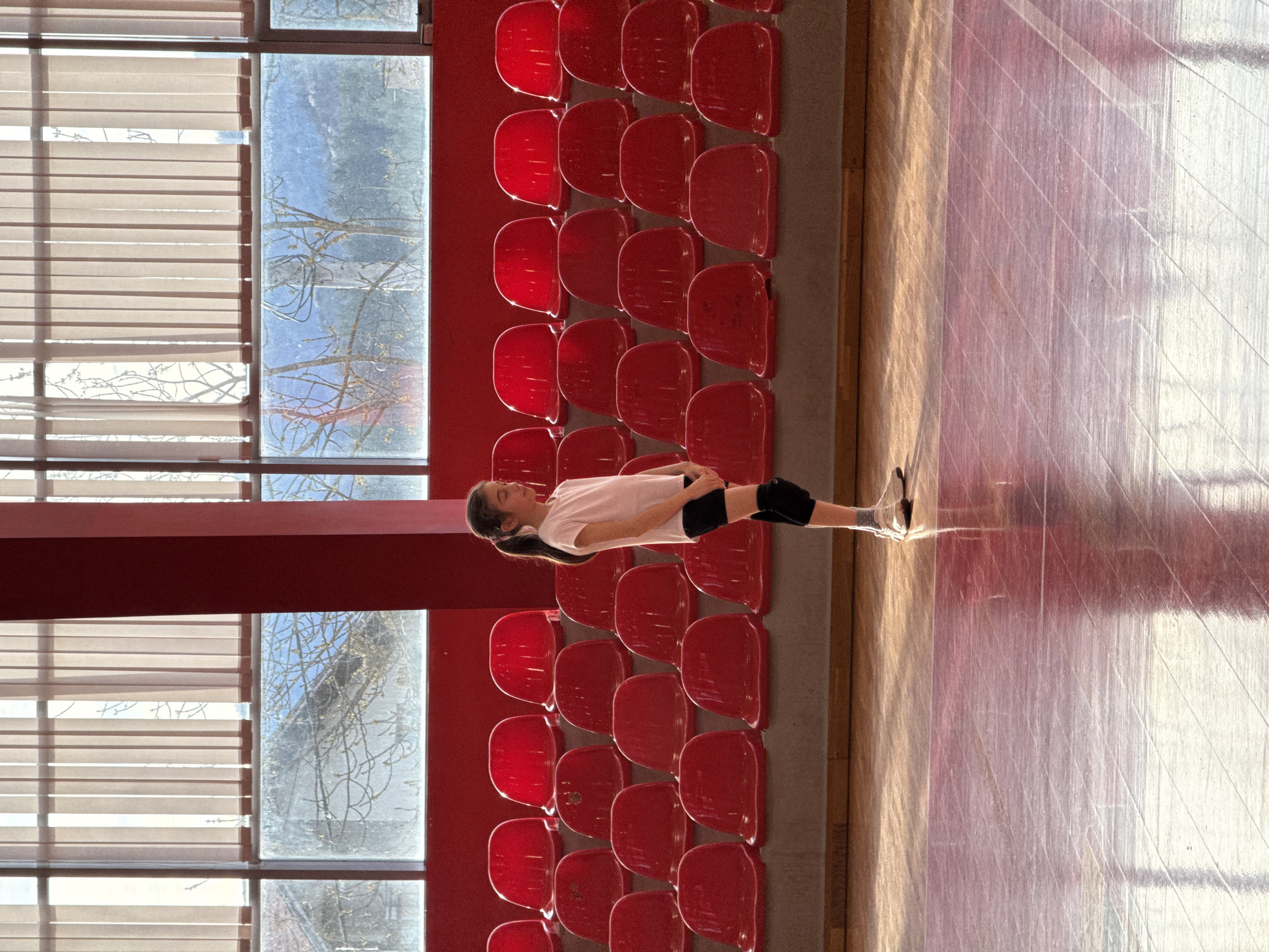 A medium shot captures a young girl stretching on a wooden floor, possibly a gymnasium, against a backdrop of red stadium seating and large windows. The girl wears a white t-shirt, black shorts, black knee pads, and grey socks. Her dark hair is pulled back into a ponytail. The stadium seating is arranged in a multi-rowed block. To the left of the seating are large windows with blinds, and a glimpse of tree branches can be seen outside, suggesting it is daytime. The wooden floor reflects light from the windows. Based on the location provided, this scene is likely occurring in Etropole, Bulgaria.