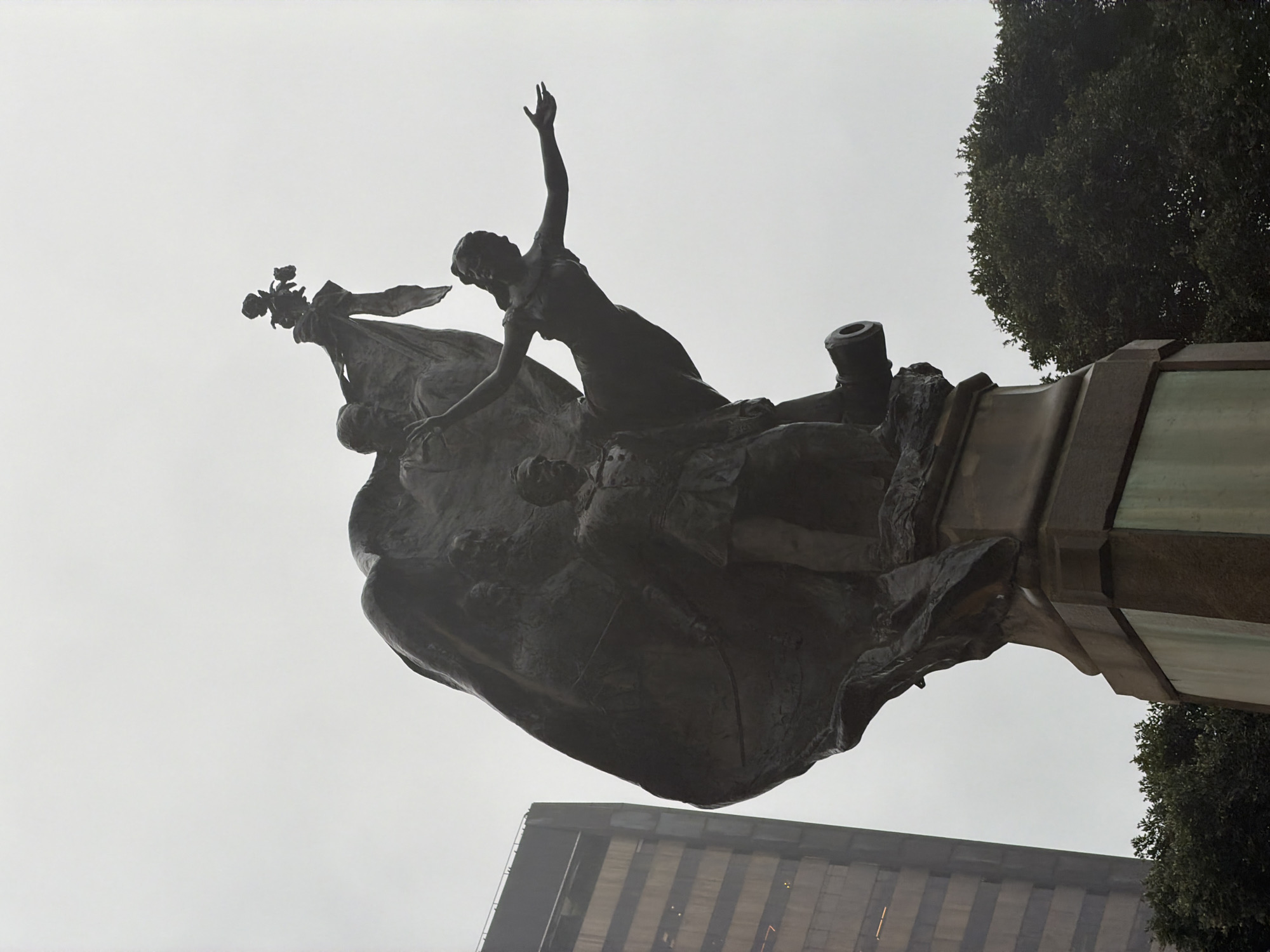 The image shows a bronze statue of a male figure in military uniform, standing on a pedestal. A draped flag flows behind him, and a female figure with outstretched arms appears to be emerging from the flag. There is also a cannon visible on the right side of the statue. In the background, a tall building with many windows and a dense green tree are visible. The statue is located at the Campo de Santana in the city of Rio de Janeiro, Brazil. The sky is overcast, indicating a cloudy day.