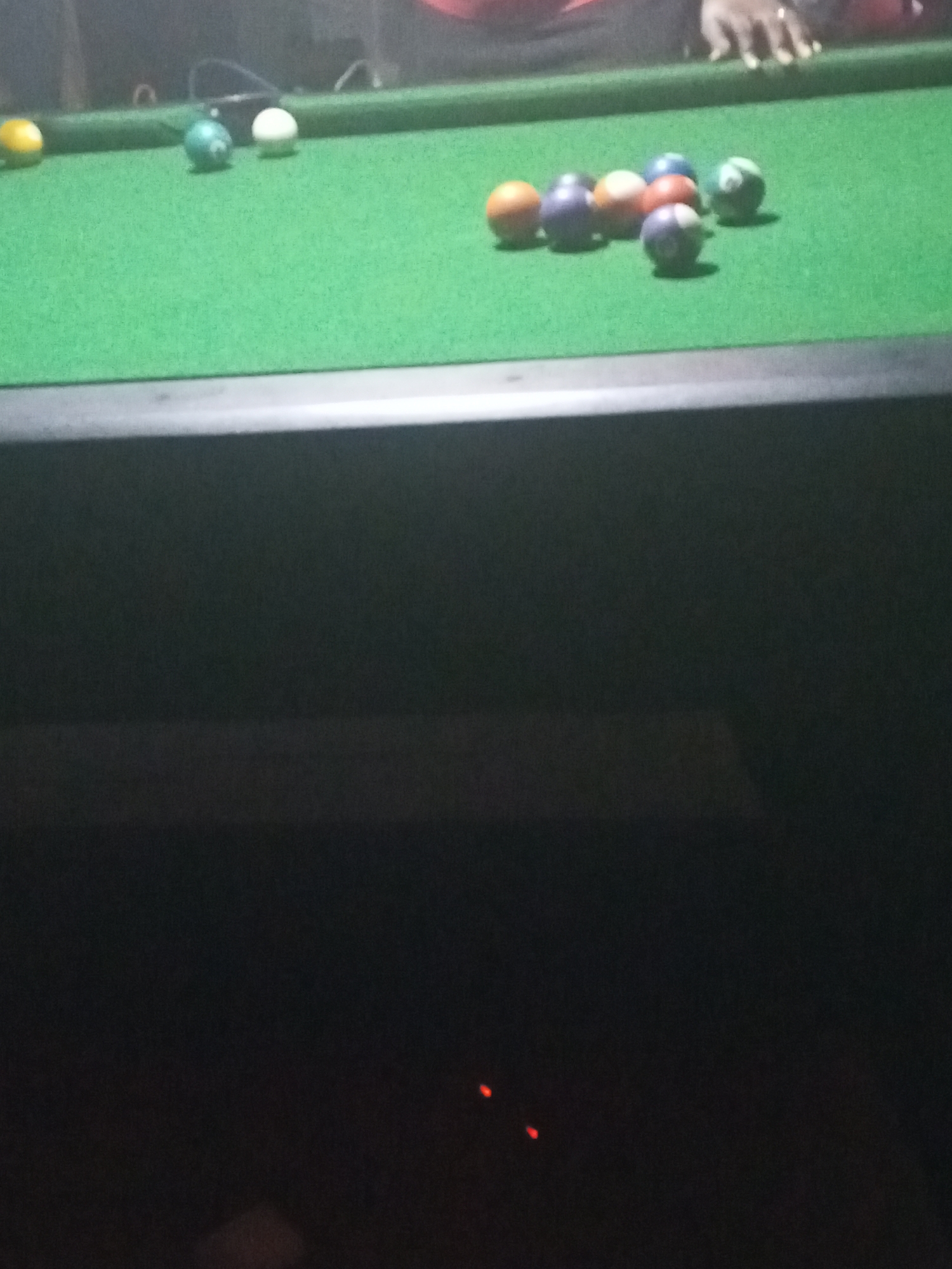 The image displays a close-up view of a green-felted pool table, likely situated indoors in a dimly lit environment. The upper portion of the frame captures the playing surface, while the lower part is obscured by the dark frame of the pool table and the indistinct, dark surroundings.
Numerous pool balls are visible on the table. On the left, a solid yellow ball, a solid teal or blue ball, and a white cue ball are present. Towards the right side of the table, a cluster of balls includes solids in orange, purple, and blue, along with a white-and-green striped ball and a white-and-purple striped ball. A human hand, with a light skin tone, is seen in the upper right quadrant of the image, resting near some of the balls on the table's edge, suggesting a game of pool is either underway or about to begin.
The lighting is low, implying the photograph was taken during the evening or night, or in a room with intentionally subdued illumination. The background is dark and out of focus, revealing no specific details about the setting beyond the immediate pool table. No text is visible within the image. The scene conveys a relaxed atmosphere typical of a recreational activity. The visual details in the image itself do not offer specific geographic cues to confirm the location as Lokoja, Nigeria, beyond the provided context.