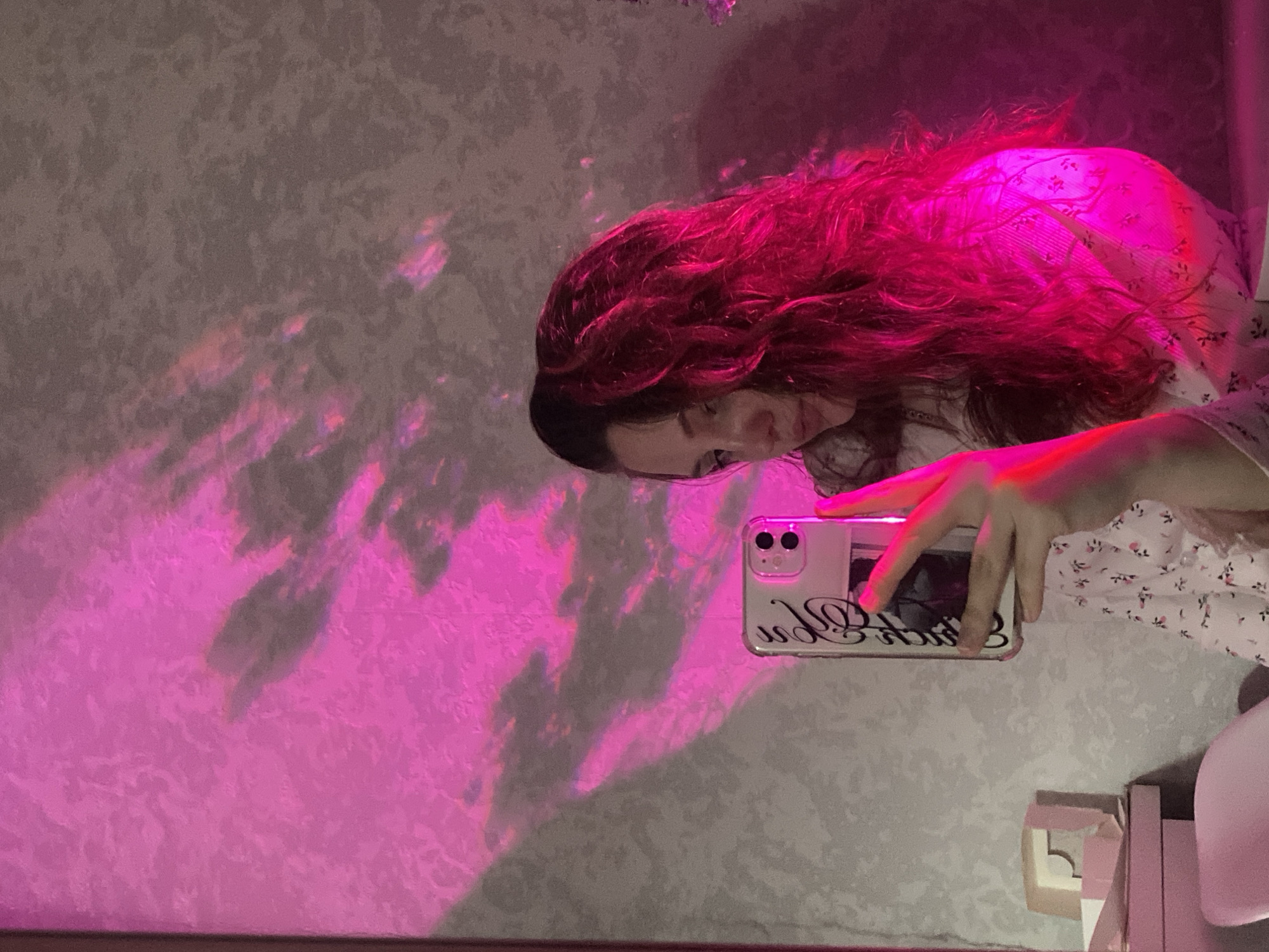 A woman with long, wavy red hair is holding a smartphone. The phone has a light-colored case with a black inscription. She is wearing a light-colored top with a floral pattern. The wall behind her is decorated with a mottled pattern and is illuminated by a pink light. The location is unknown and cannot be determined from the image. The city of Izhevsk, Russia is not visible in the image.
