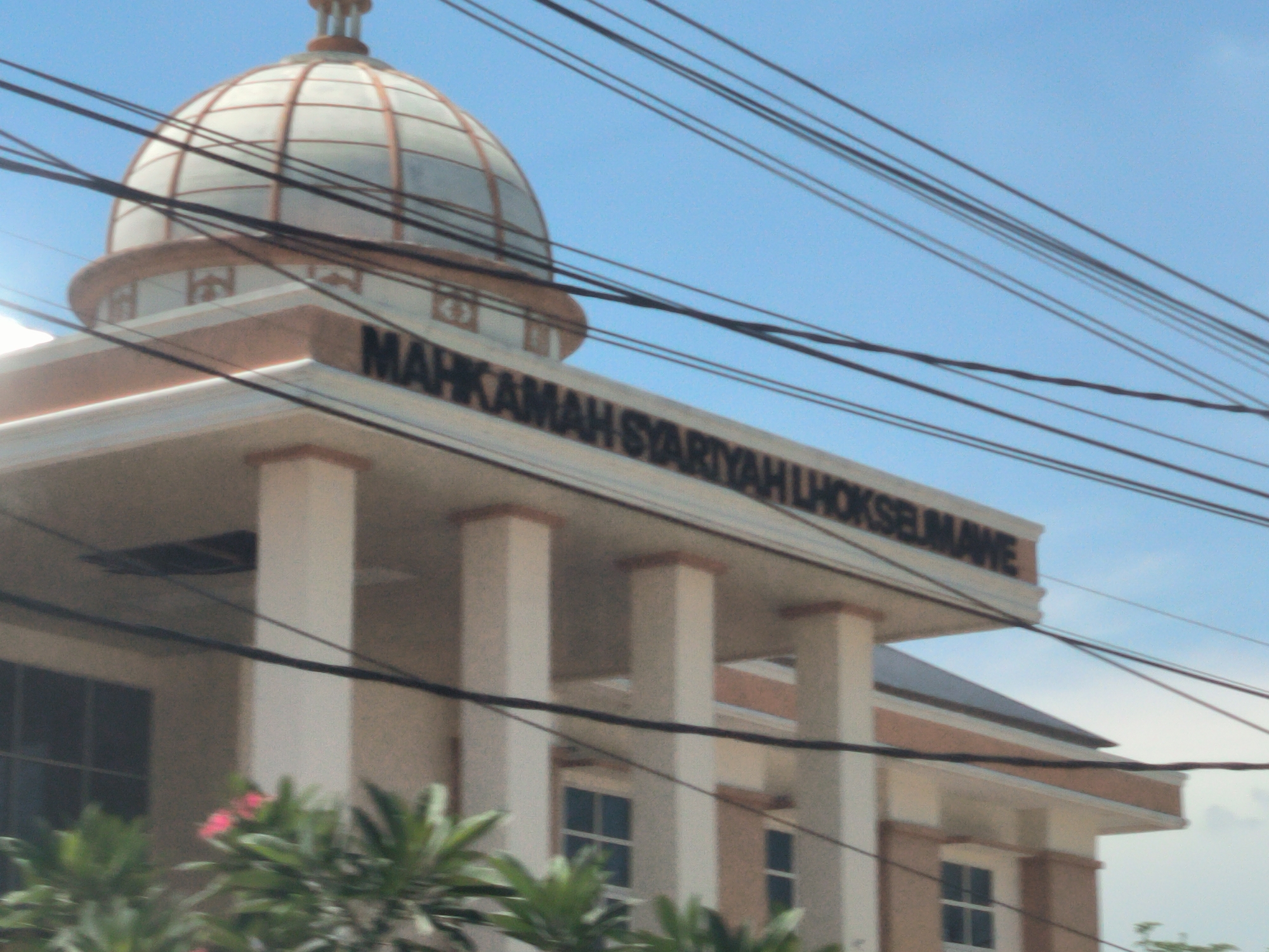 The image depicts the exterior of Mahkamah Syar'iyah Lhokseumawe, a Sharia court in Lhokseumawe, Indonesia. The building features a dome structure atop a flat roof supported by columns. The building's name is displayed in black lettering on the facade. Power lines are visible in the foreground against a blue sky. Green foliage is present at the base of the building. The building appears to be two stories high, with windows visible on both levels. The architectural style is modern with Islamic influences.