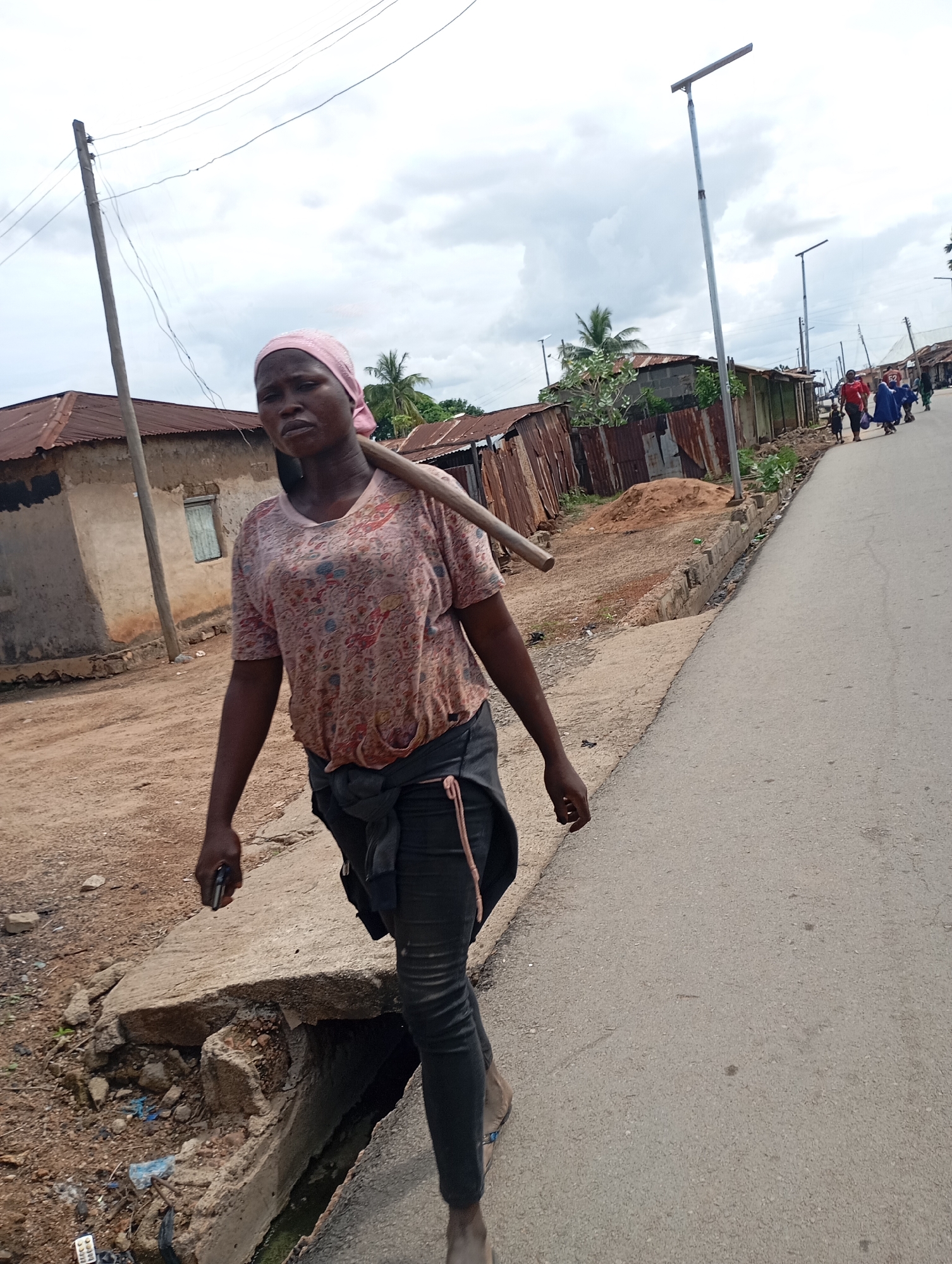 In Shendam, Nigeria, a woman walks on a dirt path next to a paved road. She wears a pink headscarf and a patterned t-shirt, with dark pants tied around her waist. A wooden pole rests on her shoulder. She holds a black object, possibly a phone, in her left hand. To her left, there are houses with corrugated iron roofs and a dirt-filled drain. Further down the road, several people, some carrying bags, walk towards the horizon. The sky is overcast with clouds.