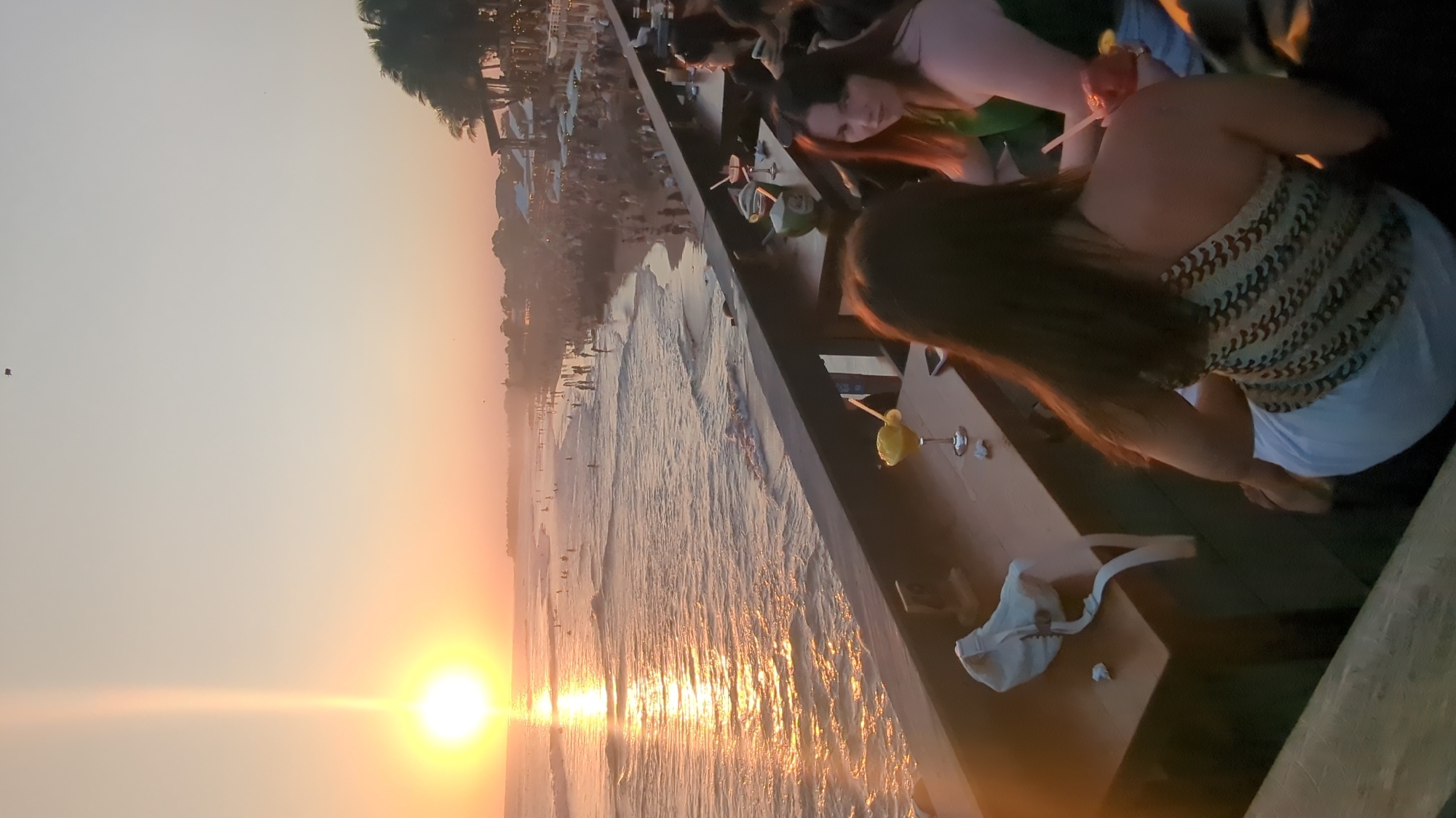 Captured at sunset, the image shows an elevated perspective over the ocean in North Kuta, Indonesia. The setting sun creates an orange glow across the sky and reflects on the water's surface. From the edge of a wooden platform, people are visible, some holding drinks. On the platform's surface are glasses, bags, and other personal items. The beach below is dotted with people and surfboards. In the background, there are buildings and palm trees.