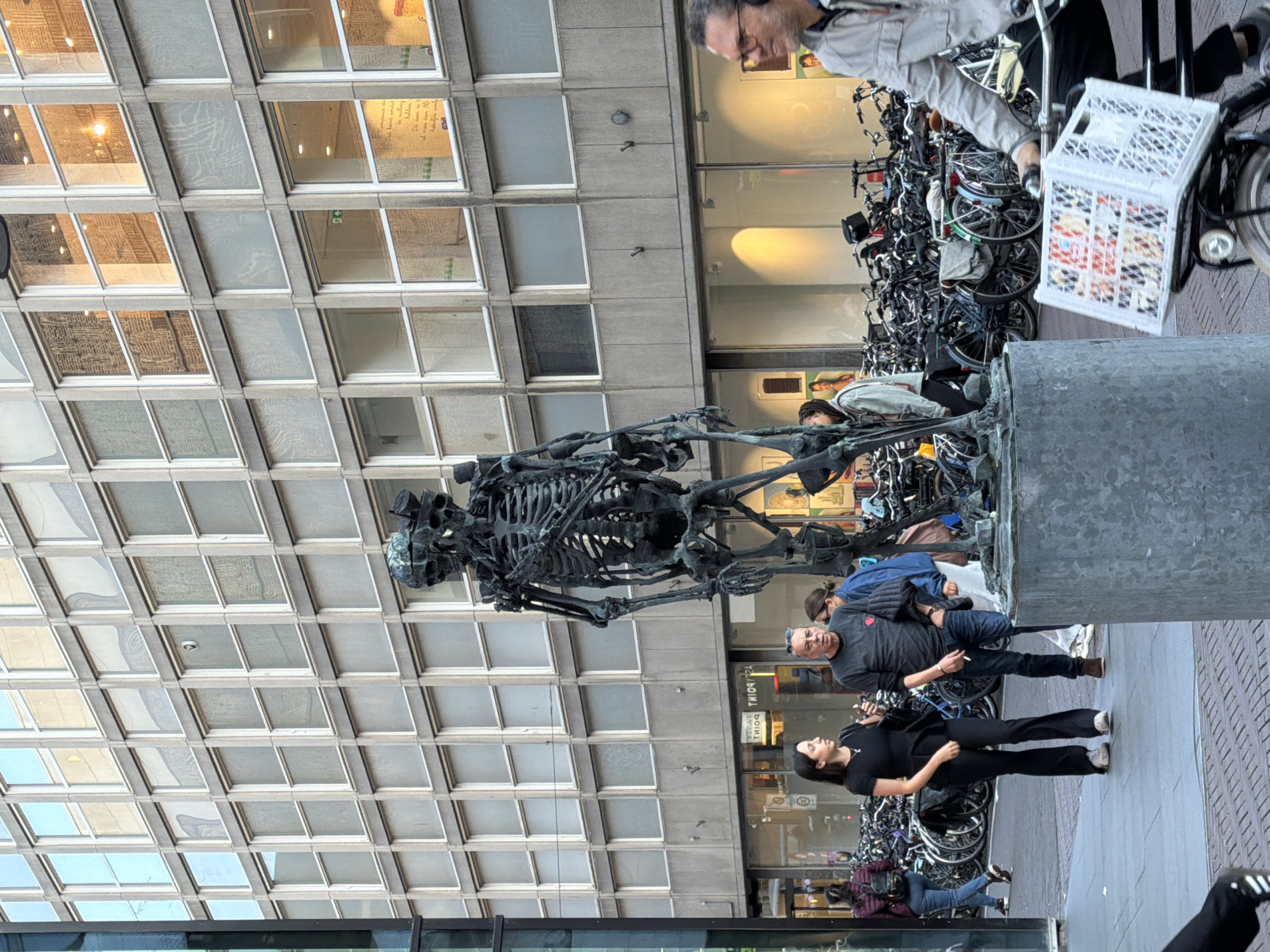 This media file captures an outdoor scene in The Hague, Netherlands, with a focus on people interacting with a large, dark sculpture of a skeleton. The sculpture is positioned in the foreground, dominating the lower portion of the frame. The background features a modern building with a facade composed of numerous square and rectangular windows, reflecting the ambient light.
Several individuals are visible in the scene. In the mid-ground, a man with glasses and a gray jacket is looking downwards, holding a cigarette. He is standing near a dense rack of bicycles. To his right and slightly behind him, a woman in a black outfit is looking to her left. Another woman, also in black, is partially visible in the lower right of the frame. Further back, near the bicycles, other individuals can be seen, one wearing a blue jacket and another partially obscured by the sculpture.
The scene appears to be set during the daytime, with natural light illuminating the area. The weather seems pleasant, as people are dressed in light to medium clothing. The presence of numerous parked bicycles suggests a public space, possibly near a commercial area or public transportation hub.
Notable details include the intricate structure of the skeleton sculpture, the organized rows of bicycles, and the geometric pattern of the building's windows. The man with the cigarette appears relaxed, while the woman in black has a pleasant expression. The overall atmosphere suggests a casual, everyday moment in an urban environment. No discernible text is clearly visible in the media file.