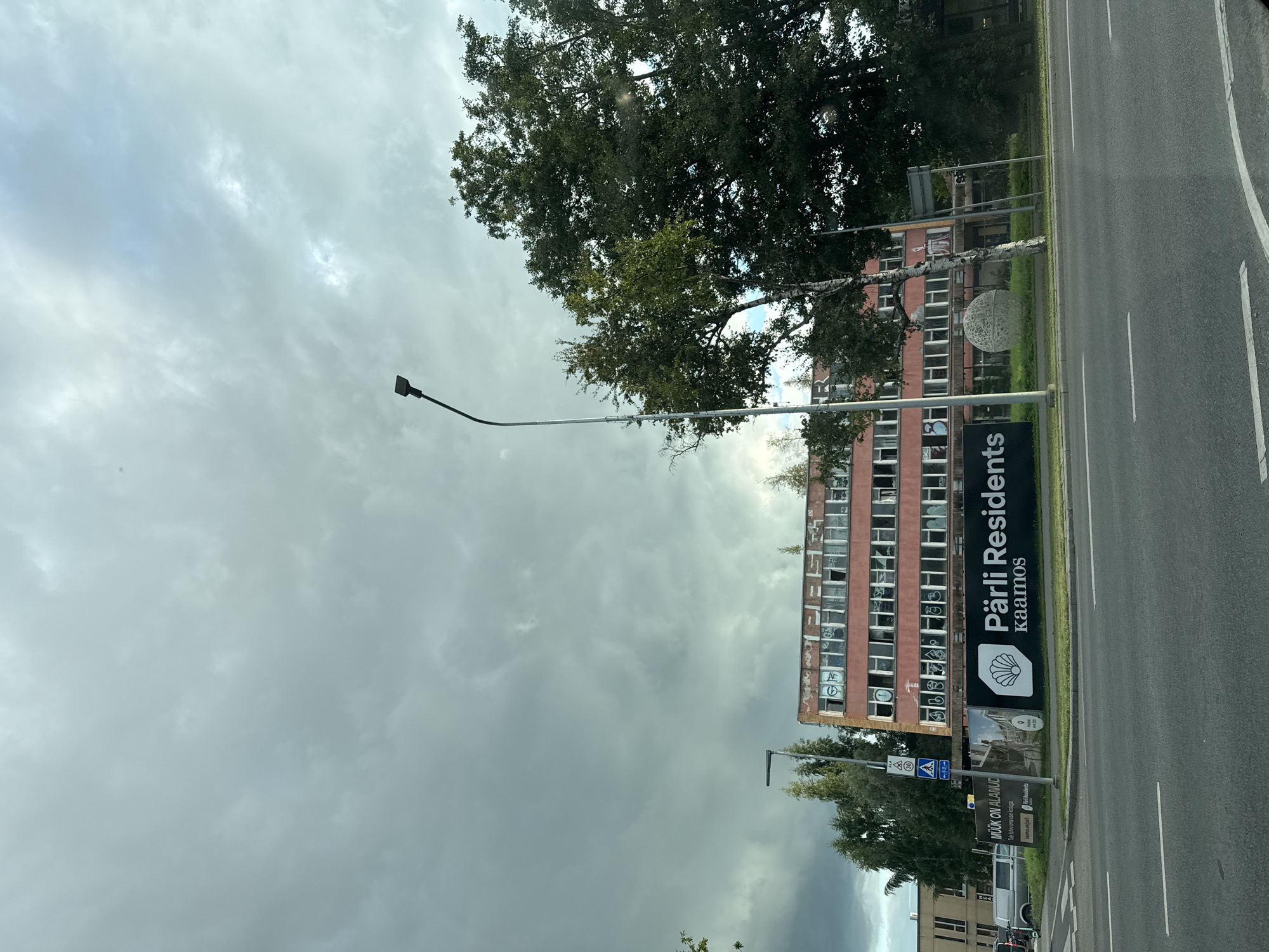 The image depicts a street view in Tallinn, Estonia, featuring a large, pink, multi-story building with many broken windows and graffiti. A dark cloud-filled sky looms overhead. In the foreground, a street with white lane markings is visible, along with a sidewalk and grass verge. To the left of the building, a billboard displays "PÄRLI RESIDENTS kaamos" with a shell logo. Next to the billboard are traffic signs and a street lamp. Trees, including a birch tree, are scattered around the building and along the street. To the far left, another building and a parked van are partially visible. The overall scene suggests a somewhat derelict urban environment under an overcast sky.