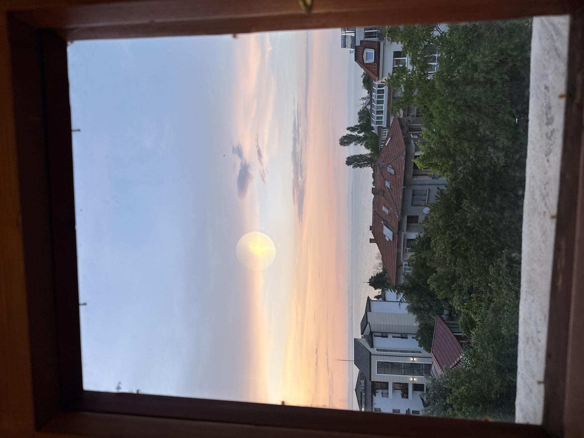 The image captures a picturesque view from inside a wooden window frame, looking out at a coastal landscape during what appears to be either sunrise or sunset. The window frame itself is dark brown, partially obscuring the top and sides of the scene.
Through the window, the sky dominates the upper portion of the exterior view, painted with soft, pastel hues of light blue, pink, and orange, typical of the golden hour. A prominent, bright, circular reflection, likely of the sun, is visible on the window glass in the upper left quadrant, creating a lens flare effect. Several wispy clouds are scattered across the sky.
Below the colorful sky, a calm body of water stretches across the horizon, separating the sky from the coastal landmass. On land, a collection of residential buildings with classic red-tiled roofs and light-colored facades are visible. These structures feature multiple windows and some balconies, reflecting a typical European coastal town architectural style. Lush green trees, some quite tall, are interspersed among the buildings, adding natural vibrancy to the urban environment. The scene suggests a tranquil moment, capturing the serene beauty of the coastline of Chernomorets, Bulgaria. There is no visible text in the image.