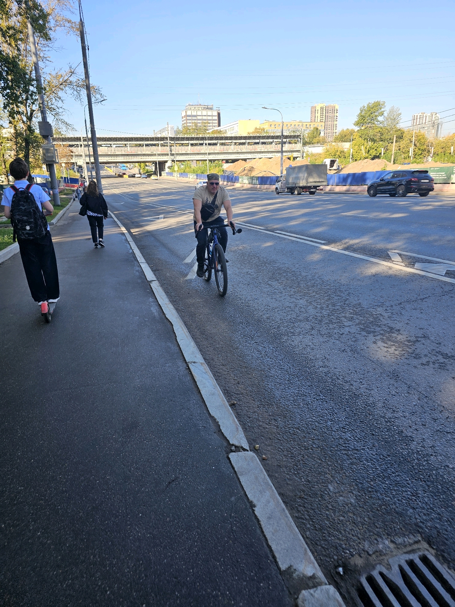 The image depicts an urban scene in Moscow, Russia, likely during the daytime with clear skies. In the foreground, a person is riding an electric scooter on a paved pedestrian walkway. Behind them, another person is walking on the same walkway. A third individual is cycling on the adjacent road. The road features multiple lanes of traffic and appears to be a busy thoroughfare.
In the middle ground, several vehicles are visible, including a truck, a black SUV, and other cars. Construction is evident in the background, with piles of dirt and blue fencing suggesting an ongoing project. Elevated structures, possibly a railway or overpass, are also present. Distant buildings, including multi-story residential complexes and commercial structures, form the city skyline. The overall environment suggests a typical urban setting with active transportation and infrastructure development.