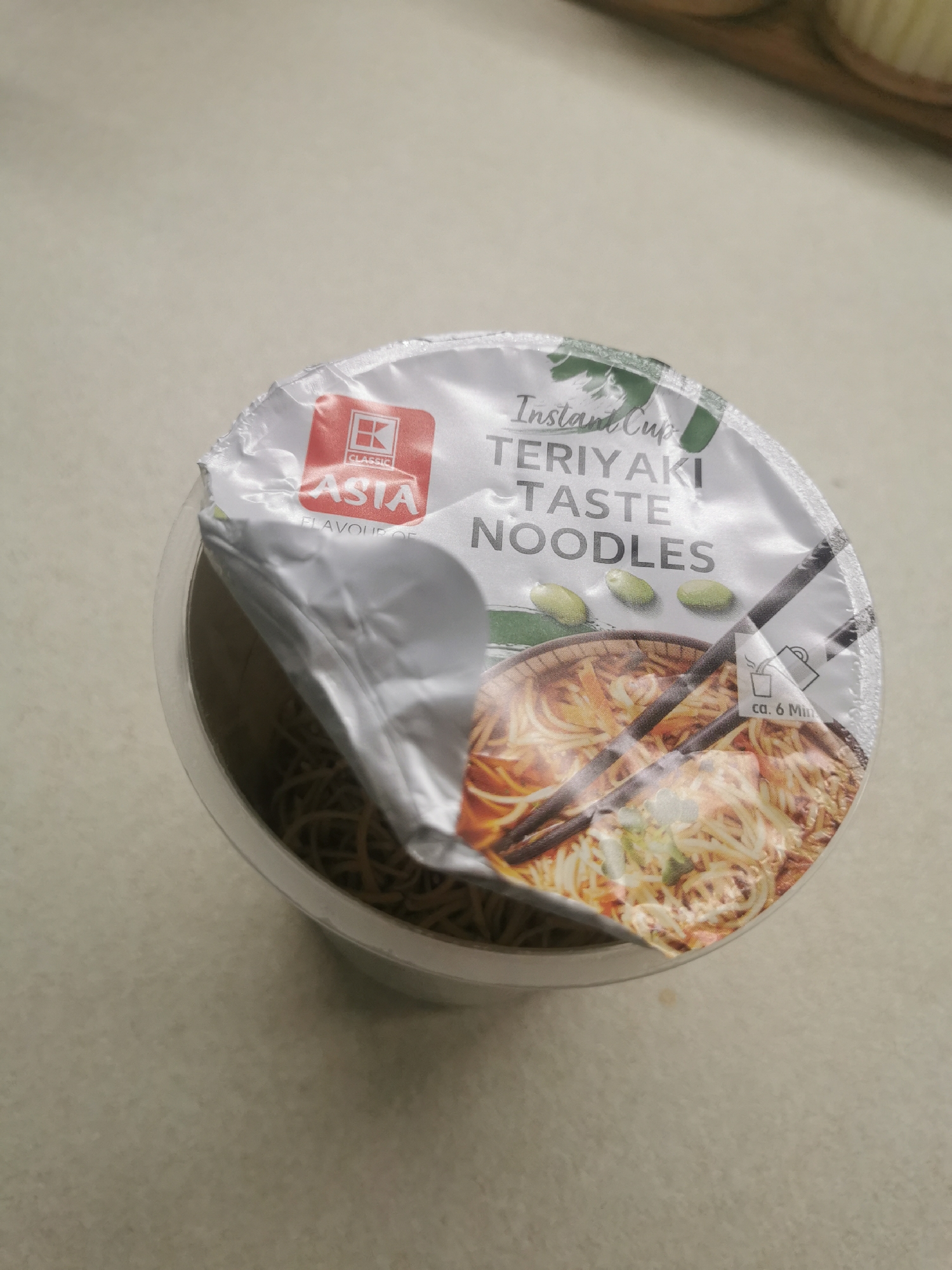 An instant noodle cup is the primary subject of this close-up, high-angle photograph. The cup, appearing white or translucent, rests on a light-colored, possibly off-white, surface, likely a kitchen counter or table indoors. The lid of the instant noodle cup is partially peeled back, revealing dry noodles inside the container.
The lid is white with green design elements and black text. Prominently displayed text on the lid reads "INSTANT CUP", "TERIYAKI TASTE NOODLES". A red square logo with a white "K" and the word "CLASSIC" above "ASTA" is visible on the left side of the lid, along with the words "FLAVOUR OF". An appetizing image of cooked noodles in a bowl with chopsticks is printed on the lid, along with three green, bean-like vegetables. A small icon indicating adding water and a text "ca. 6 Min" (approximately 6 minutes) suggests preparation instructions. The partial peeling of the lid implies the noodles are either about to be prepared or are in the process of preparation. No people are visible. The lighting is bright and even, suggesting daytime. The background is a plain, out-of-focus surface. While the context is Blagoevgrad, Bulgaria, no specific visual elements within the image tie it to that location.