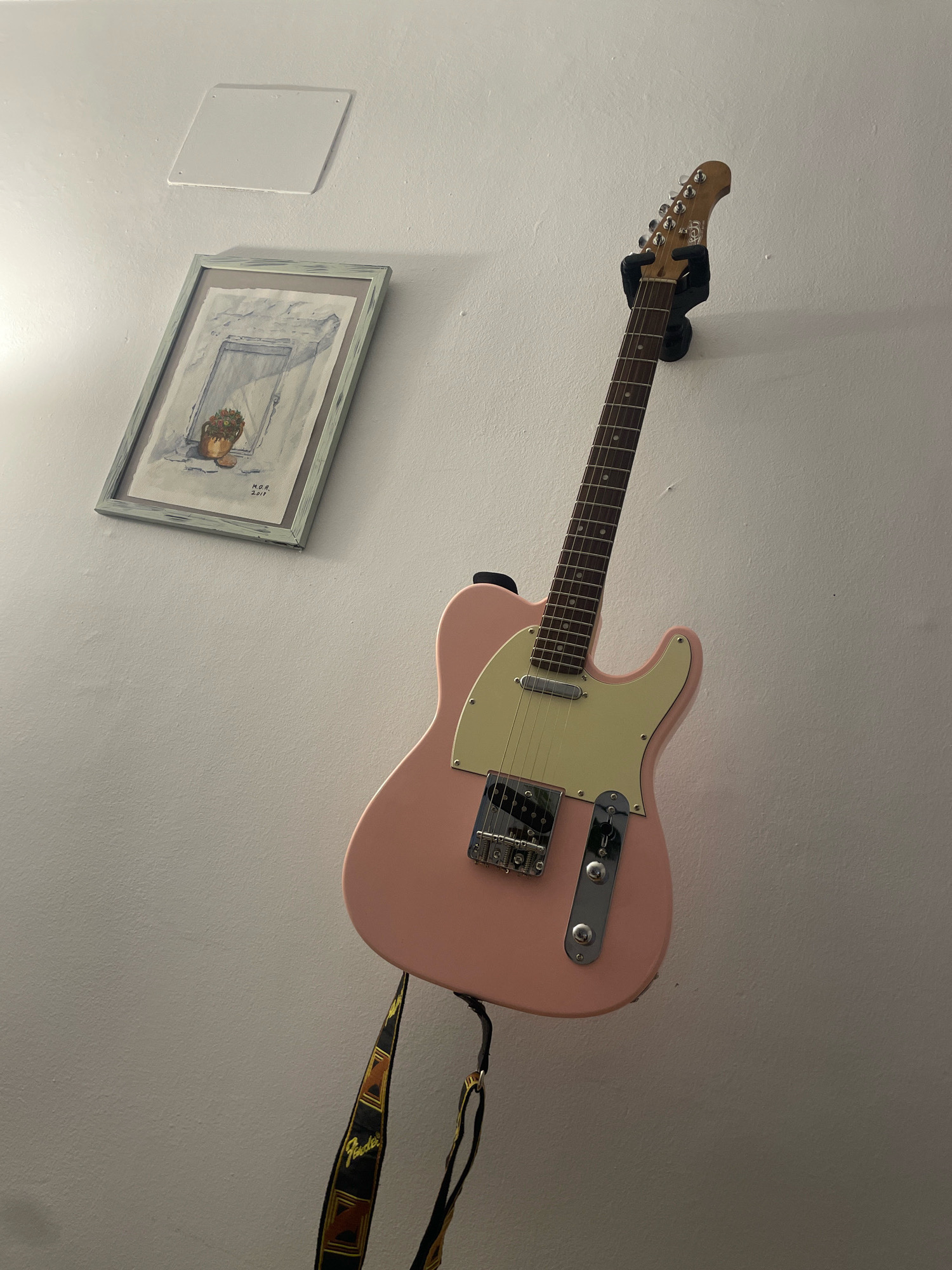 A pink electric guitar is mounted on a wall, with a black guitar strap hanging from it. To the left of the guitar is a framed watercolor painting of a doorway with a pot of flowers on a windowsill. Above the painting is a white, square electrical cover plate. The scene appears to be in a domestic setting in the city of Atarfe, Spain. The lighting is dim, casting shadows on the white wall.