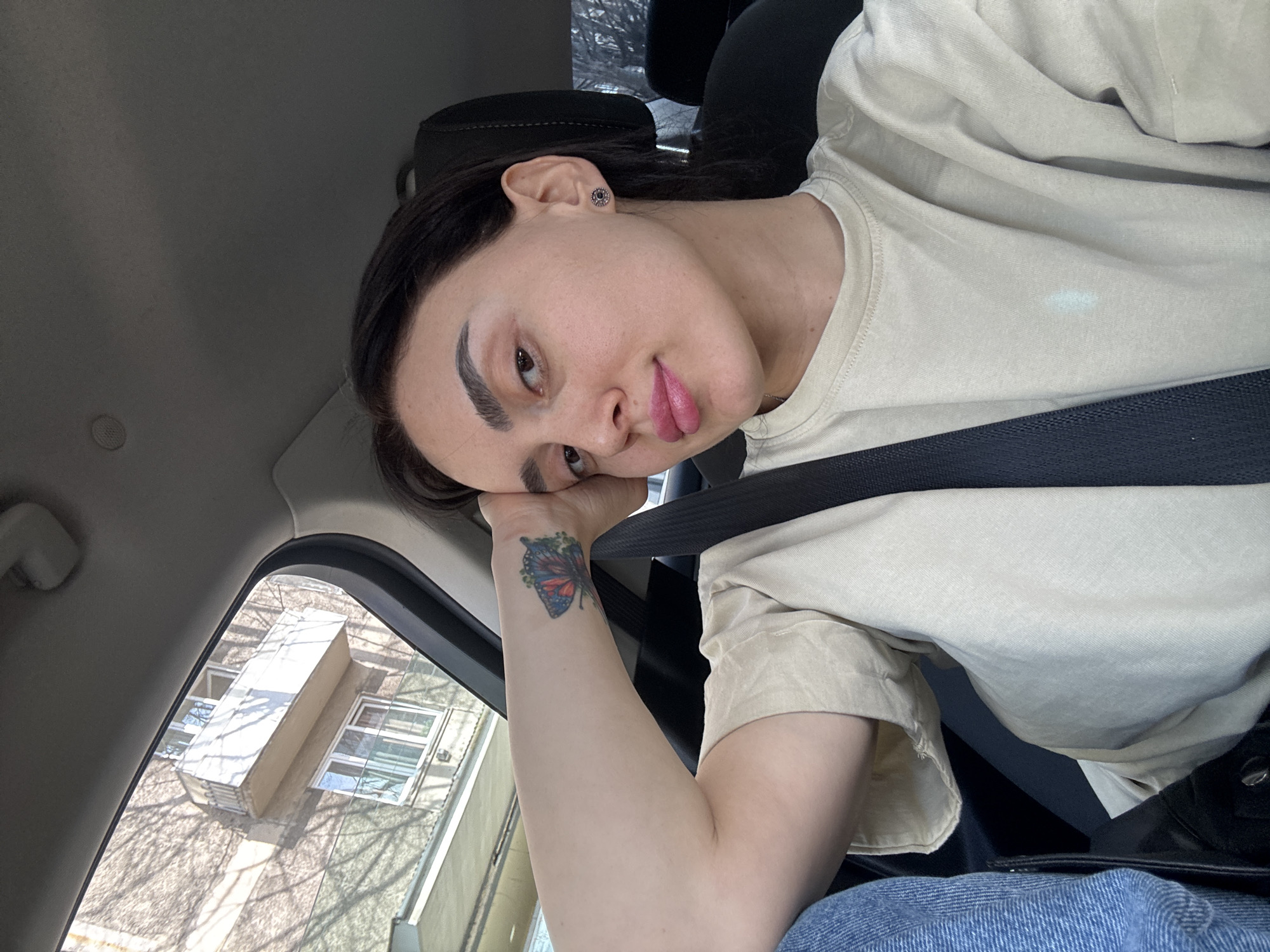 A person is shown in a car, wearing a beige t-shirt and a seatbelt. The person is leaning their head on their left hand, which has a butterfly tattoo on the wrist. Outside the car window is a building. Based on the architecture observed through the car window, it may be presumed that this image was taken in the city of Tbilisi, Georgia, though this cannot be definitively confirmed.