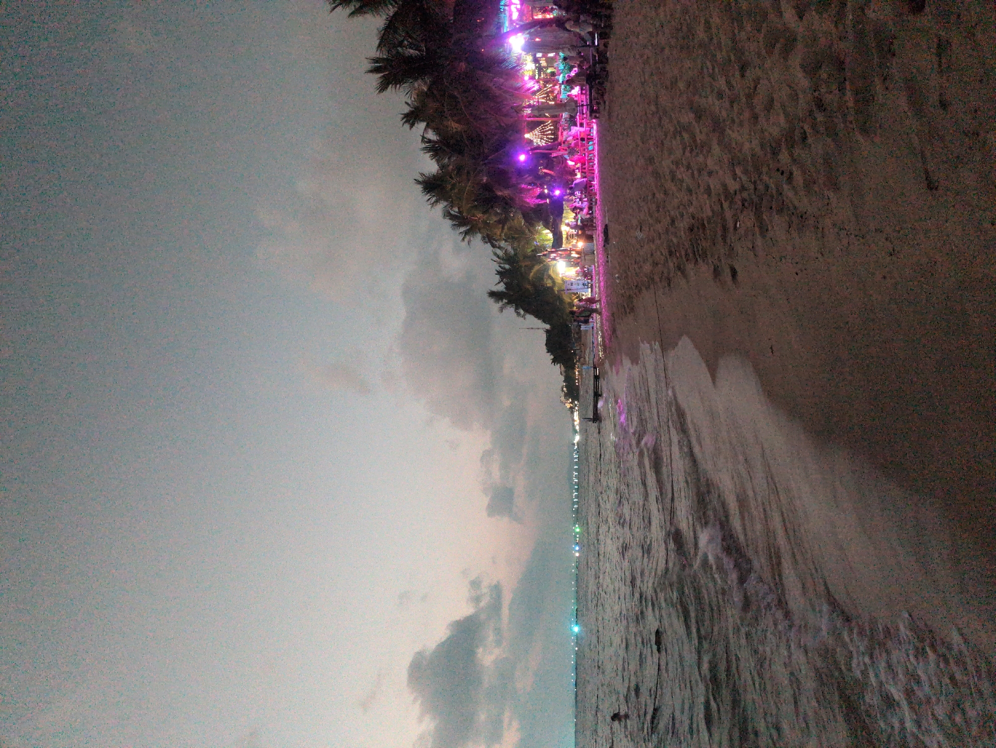 The image shows a beach scene in Phú Quốc, Vietnam at dusk or night. The view is oriented sideways, with the sky and ocean on the left, and the beach on the right. The sky is overcast with grey clouds. The ocean is dark, with a line of lights visible along the horizon, likely from boats or the coastline. The beach is sandy and slopes upward toward a line of palm trees, which are decorated with colorful lights, including pink and purple. A crowd of people can be seen among the palm trees and the illuminated area, suggesting a beachside bar or restaurant.
