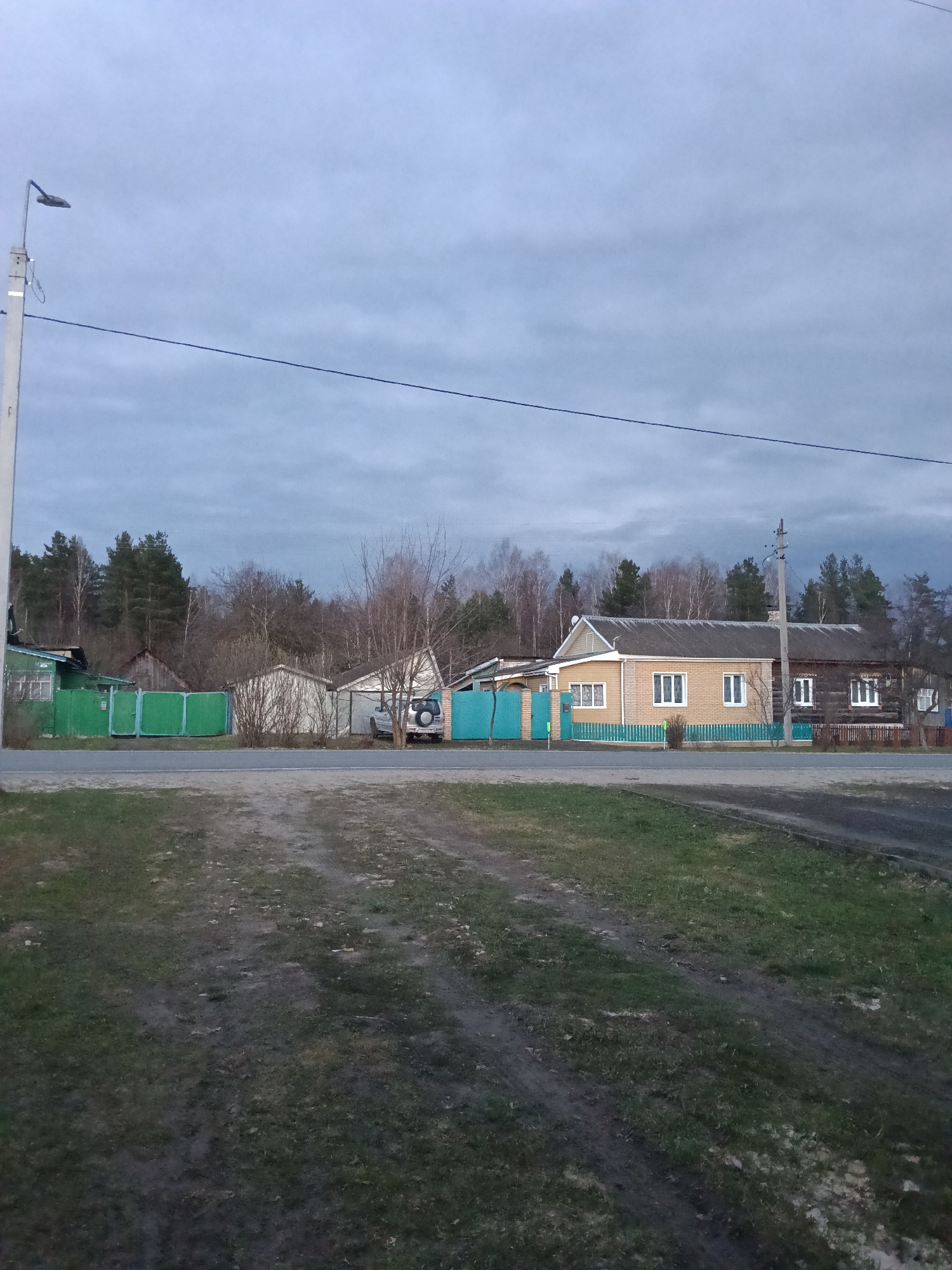 The image presents a static, rural street scene in Красный Маяк, Russia, characterized by an overcast sky.
In the foreground, a wide, unpaved area functions as a path or track, featuring visible tire marks and patches of sparse, dull green grass and bare soil, suggesting damp conditions. This area leads to a narrow asphalt road that traverses the midground horizontally.
Beyond the road, several houses line the street, each with a distinct fence. On the far left, a house with green fencing is partially obscured by a utility pole topped with a street lamp. Further right, an SUV, possibly a Suzuki Grand Vitara judging by the rear-mounted spare tire, is parked near a lighter-colored building mostly hidden by bare trees and a turquoise fence. The most prominent structure is a combined house on the right: one section is made of light yellow brick or siding with white-framed windows, seamlessly joined to an older, dark wooden log house, also with white-framed windows. Both sections share a grey roof and are fronted by a bright turquoise picket fence. Another utility pole stands near this house. The trees in the midground, mostly deciduous and bare, intersperse with evergreen pines.
The background features a dense forest, consisting of both tall pine trees and bare deciduous trees, indicating a season like late autumn, winter, or early spring. The sky is entirely covered with thick, grey clouds, casting a soft, diffused light over the landscape.
No people are visible in the scene, and there is no discernible activity taking place. The overall impression is one of quiet, unassuming village life under a subdued light. There is no visible text on any objects or structures.