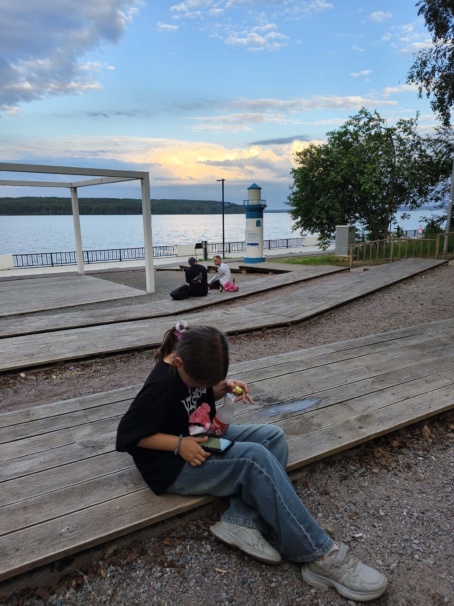 The image captures a serene outdoor scene at a waterfront park in Chaikovsky, Russia. In the foreground, a young girl with dark hair tied in a ponytail is seated on a wooden tiered seating area, engrossed in her smartphone. She is wearing a black t-shirt and blue jeans, with light-colored sneakers. Next to her, a plastic bottle is visible.
Further back, two individuals are seated on the wooden planks, appearing to be conversing. One is wearing a black t-shirt and the other a white long-sleeved shirt. A backpack is placed beside the person in the black t-shirt.
The background features a tranquil body of water, likely the Kama River, which is a prominent geographical feature of Chaikovsky. Along the waterfront, a black metal fence runs parallel to the wooden promenade. A white rectangular structure with a flat roof stands near the water's edge. Further along, a decorative lighthouse-shaped structure with blue accents is visible. Trees, including a large one on the right side of the frame, provide shade and greenery. The sky is a mix of blue with scattered clouds, and the soft lighting suggests it is either early morning or late afternoon. The overall atmosphere is one of peaceful leisure.
