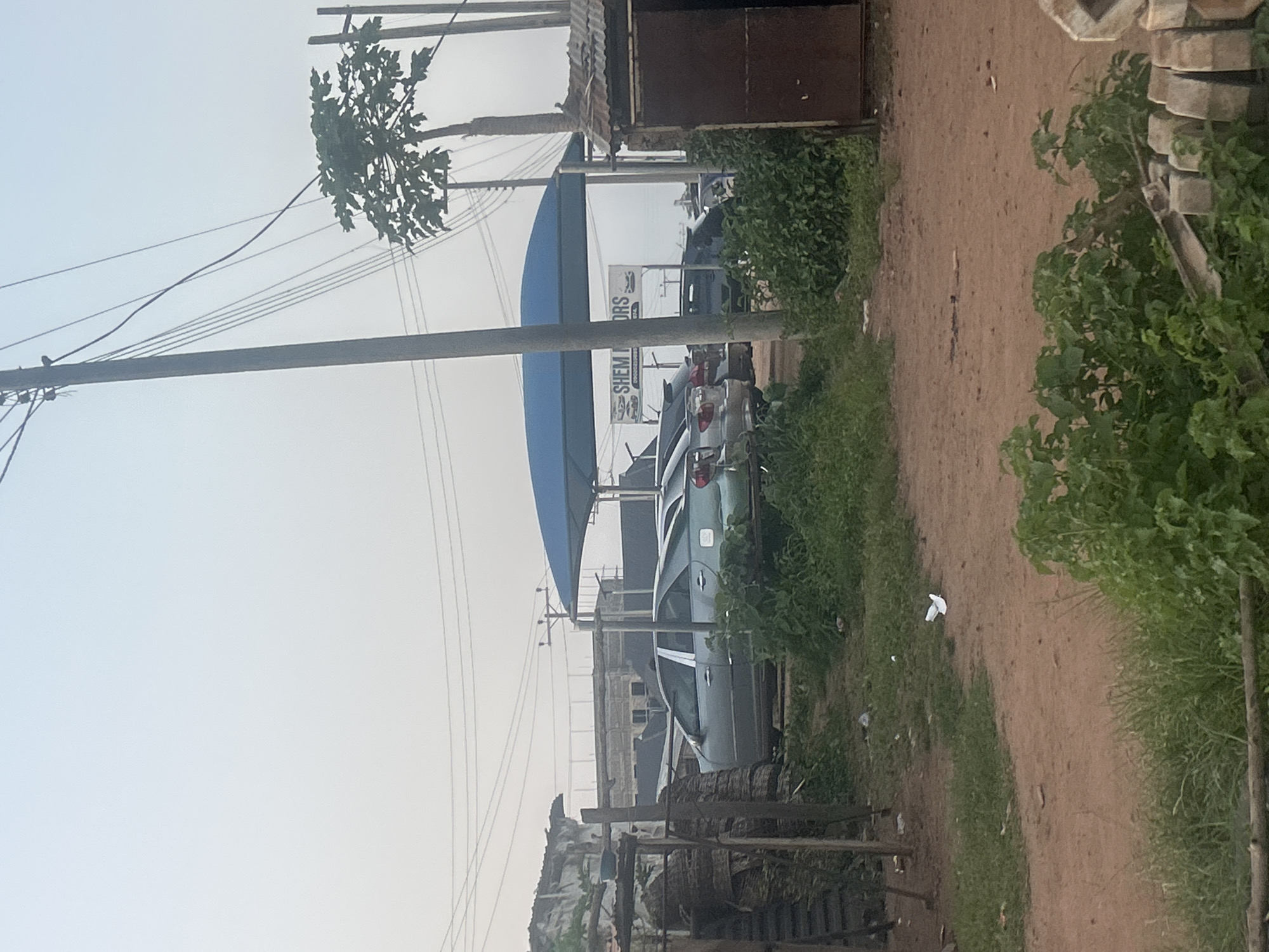 The image depicts an outdoor scene, likely a car lot, in Ogbomosho, Nigeria. Several cars are parked under a blue canopy, some of which are for sale. A sign that reads "SHEM MOTORS" is visible under the canopy. In the foreground, there is a dirt path with scattered debris, and overgrown green vegetation. To the right of the frame, a small, makeshift building constructed from corrugated metal stands. Power lines and utility poles crisscross the sky. The overall environment suggests a somewhat unkempt and utilitarian setting. There are no people visible in the image. The sky is overcast, indicating a hazy or cloudy day.