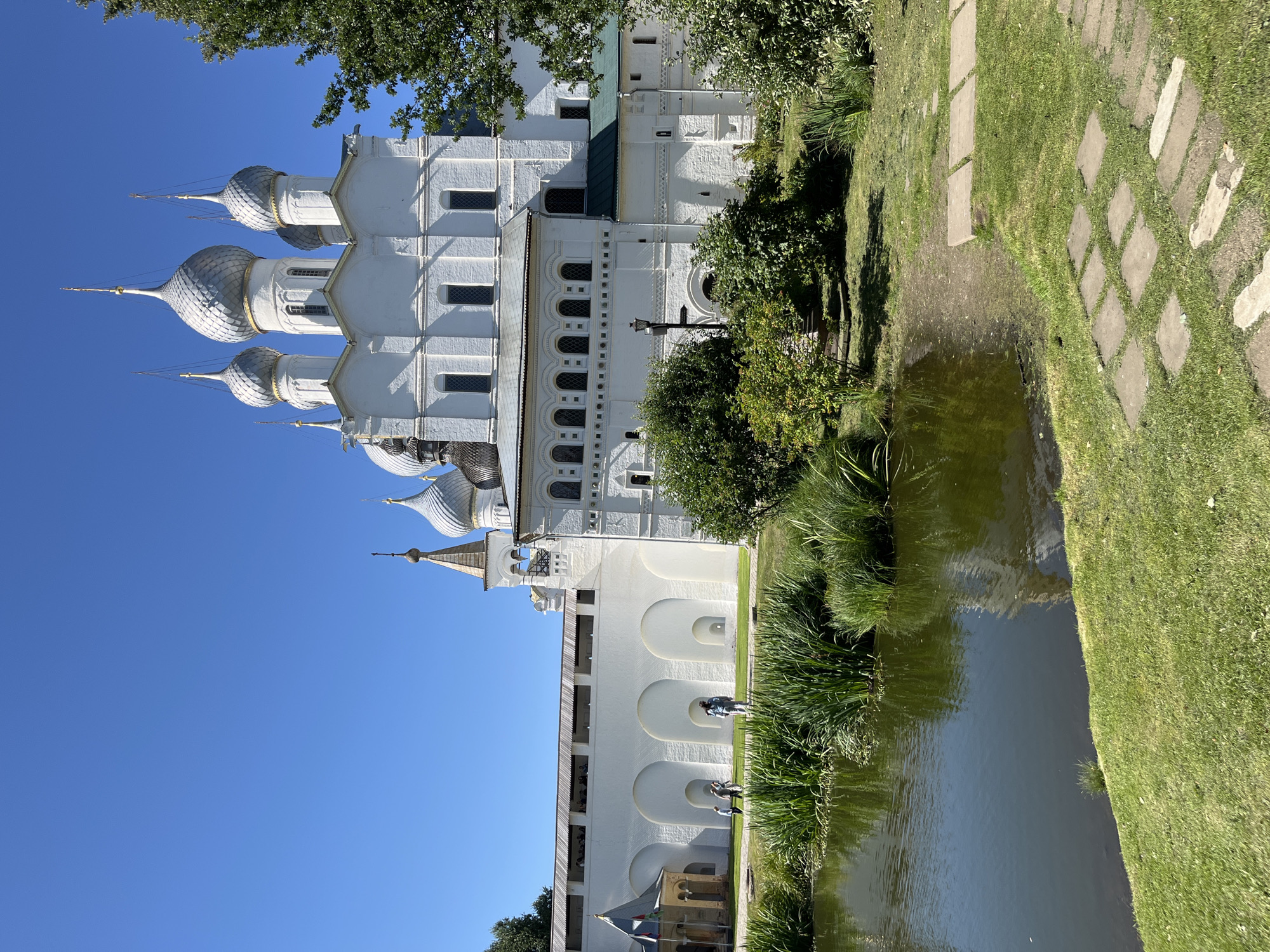 The image shows a bright, sunny day in Ново-Никольское, Russia, with a clear blue sky dominating the upper portion of the frame. In the background, a large, white, multi-domed church with silver onion domes stands prominently against the sky. The church has intricate architectural details, including arched windows and decorative cornices. To the left of the church, a white, fortified wall with arched openings stretches across the landscape. Several people are visible walking on the grounds near the wall. In the foreground, a tranquil pond with lush green reeds growing along its edge reflects the surrounding scenery. The pond is bordered by a neatly mowed green lawn, with stepping stones laid out in a pathway. Trees and bushes are scattered throughout the scene, adding to the natural beauty of the location.