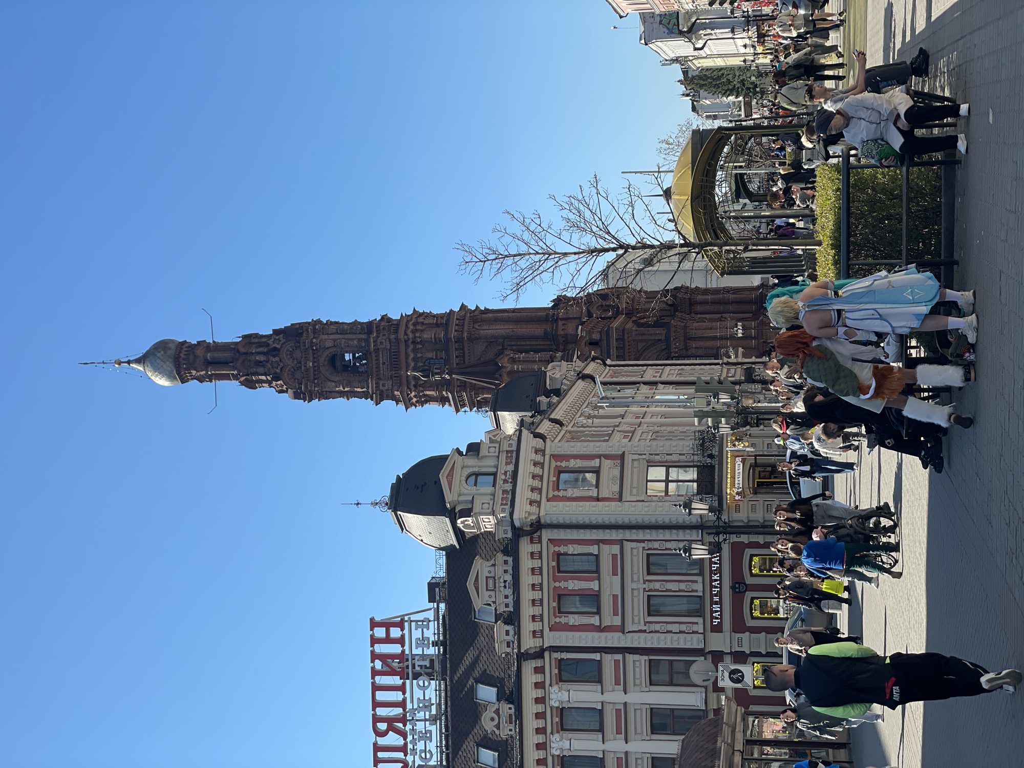 The image depicts a street scene in Kazan, Russia. The Suyumbike Tower is visible against a clear blue sky. A tall building with white and red trim is also present, adorned with a sign in Cyrillic script. People are walking and standing on the sidewalk, some appearing to be in costume. There are benches and a decorative metal structure along the street. The lighting suggests it is a sunny day.