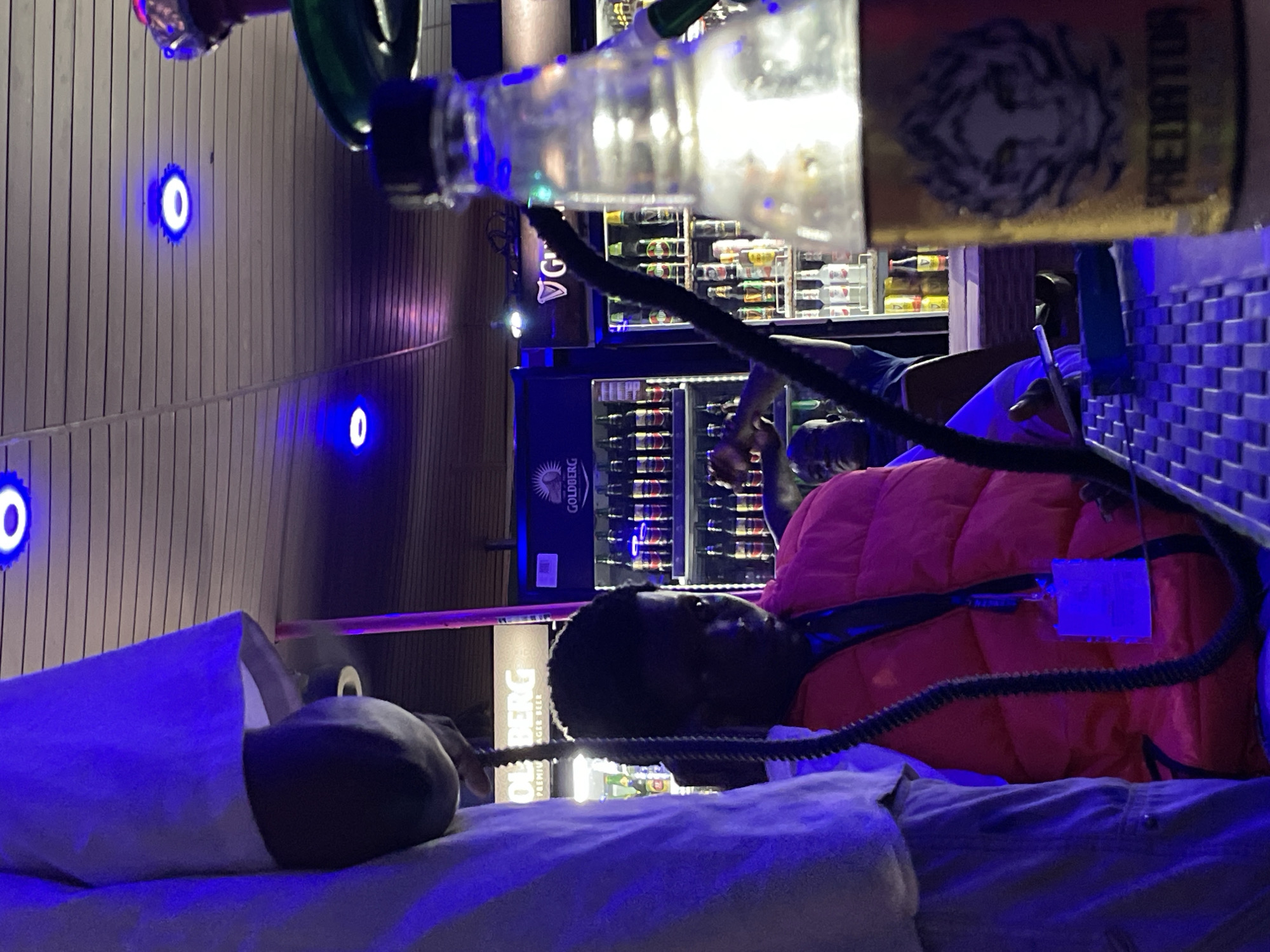 The image depicts a dimly lit interior, possibly a bar or lounge, in Ado, Nigeria, bathed in blue and purple neon lighting. In the foreground, a bottle of "Predator" energy drink is prominently displayed, its label featuring a tiger's head. A black hookah hose extends from the bottle towards the center of the frame. To the left, a person wearing a white shirt stands partially visible, their body angled away from the camera. In the mid-ground, a young person with dark hair, wearing a bright pink puffer jacket, is seated and looking downwards, holding a smartphone. They have a blue lanyard with a badge attached to their jacket. Behind them, refrigerators stocked with various beverages, including "Goldberg" beer, are visible. The ceiling is made of horizontal wooden planks with circular blue lights embedded in it. Another person, partially obscured, can be seen behind the refrigerator. The overall atmosphere appears to be casual and social.