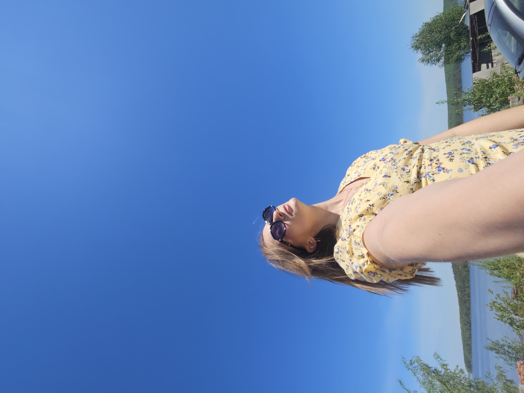 A woman with brown hair and sunglasses wears a yellow floral dress. She is positioned in front of a bright blue sky and part of a body of water surrounded by trees. A car and part of a building are visible in the upper right corner. The photograph was taken in Chaikovsky, Russia.