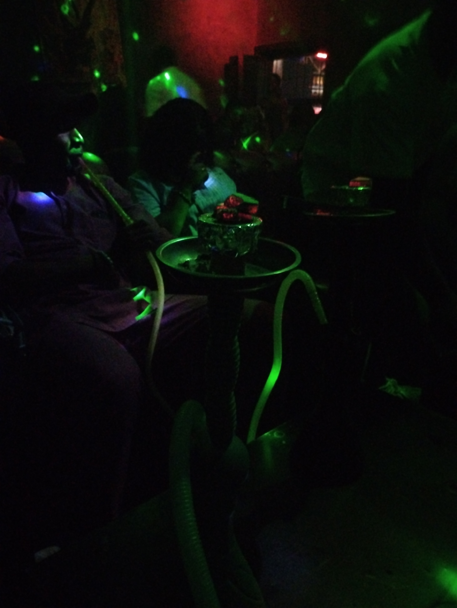 In a dimly lit interior in Gombe, Nigeria, two individuals are seated beside tables featuring hookahs. One person holds a hookah pipe to their mouth. The environment is illuminated by green and red lights.