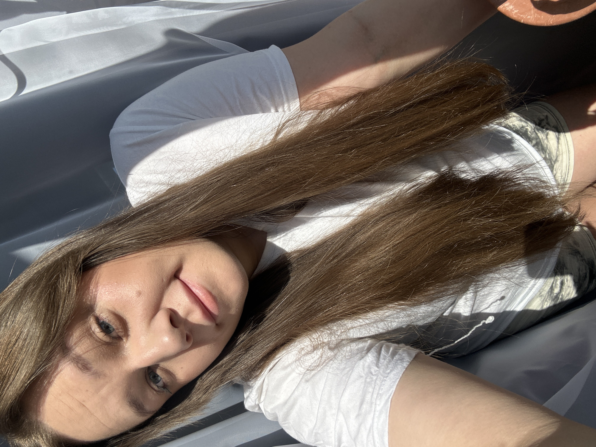 A woman with long brown hair is lying on a gray surface, possibly a bed or sofa, in a brightly lit area, most likely indoors. She is wearing a white t-shirt and shorts. Her head is tilted towards the camera, and she is looking directly at the viewer with a slight smile. The background is out of focus, but there are hints of light and shadow that suggest a window or other light source. A potted plant is in the periphery of the frame. The location is in Ольховка, Russia.