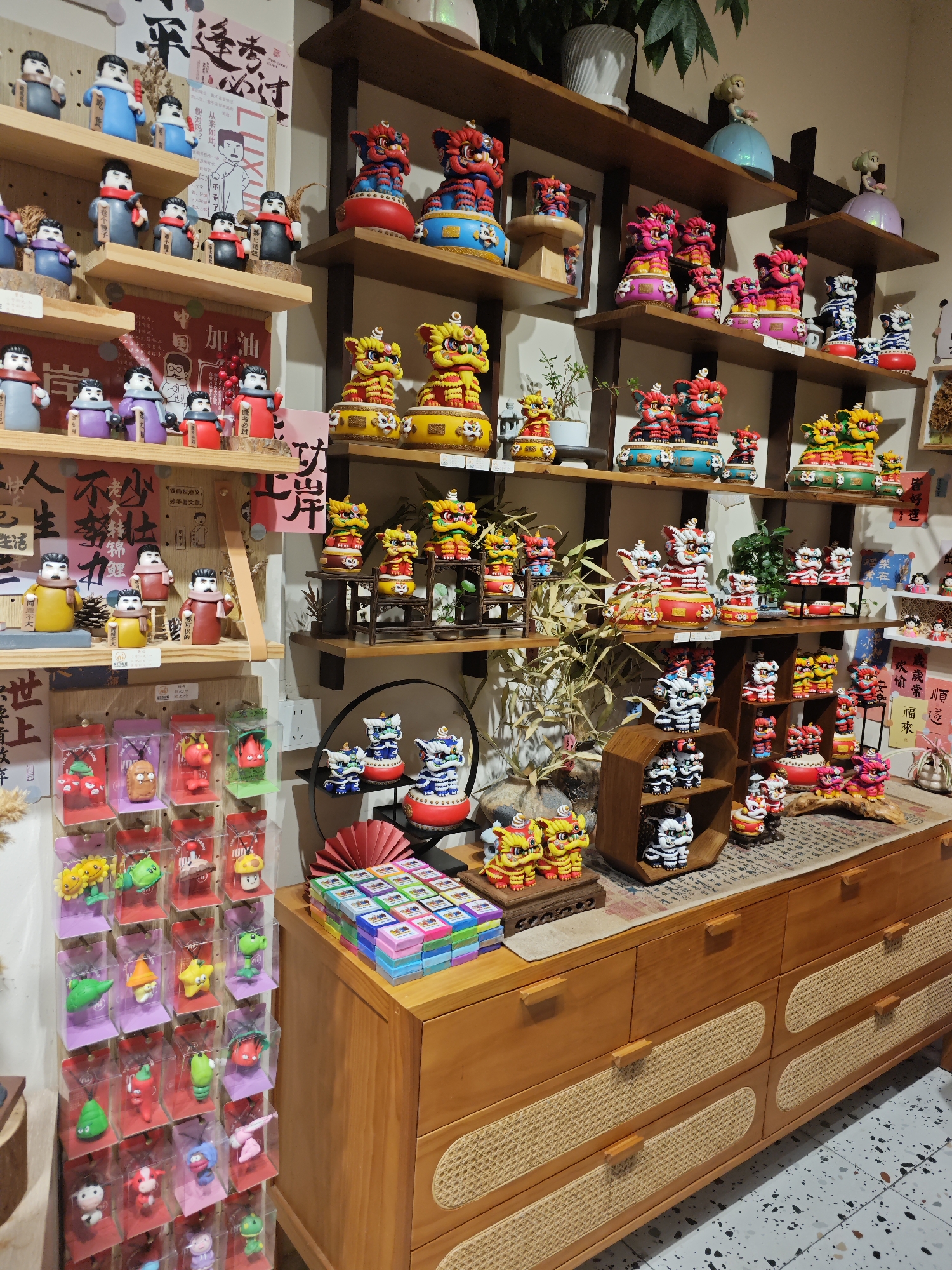 The image shows a store display in Shapingba District, China. Shelves are stocked with colorful figurines, including traditional Chinese lion dances, and dolls. A wooden cabinet with woven drawer fronts sits below, topped with more figurines and boxed items. Packaged keychains with cartoon characters are hanging at the left. Chinese calligraphy scrolls are also visible among the shelves.