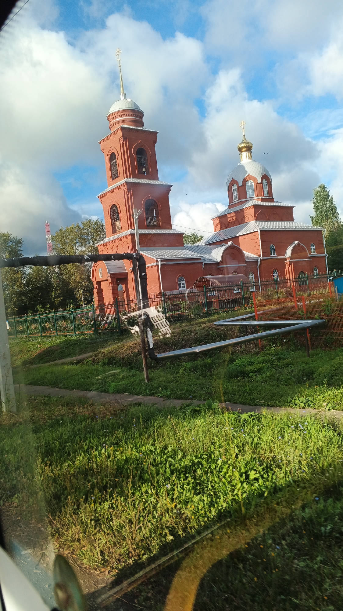 The image shows a red brick church with a tall bell tower and a dome, located in the village of Kiyasovo, Russia. The church is a traditional Orthodox Christian building with arched windows and a cross on top of the dome and bell tower. The church is surrounded by a green metal fence and lush green grass. In the background, there are trees and a partly cloudy blue sky. A cellular tower is visible to the left of the church. There are no people visible in the image. The foreground shows a road or path and some pipes or conduits running along the ground. The image appears to be taken from a moving vehicle, as evidenced by the reflection on the lower left portion of the image.
