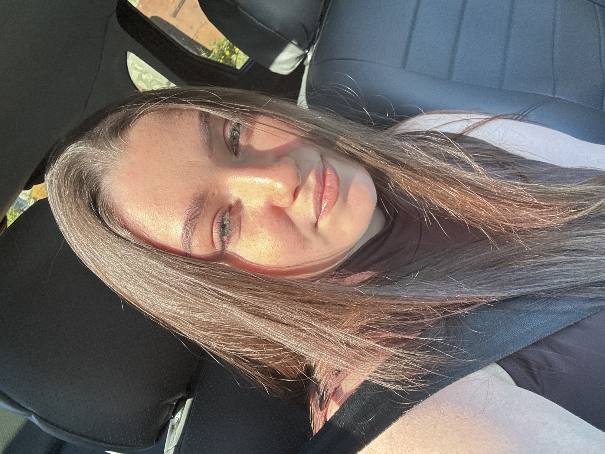 A fair-skinned woman with long, light brown hair is sitting in a car. She is wearing a seatbelt. The car's interior is black. The image is taken in bright sunlight. It is impossible to confirm the location as Гамово, Russia from the given image.