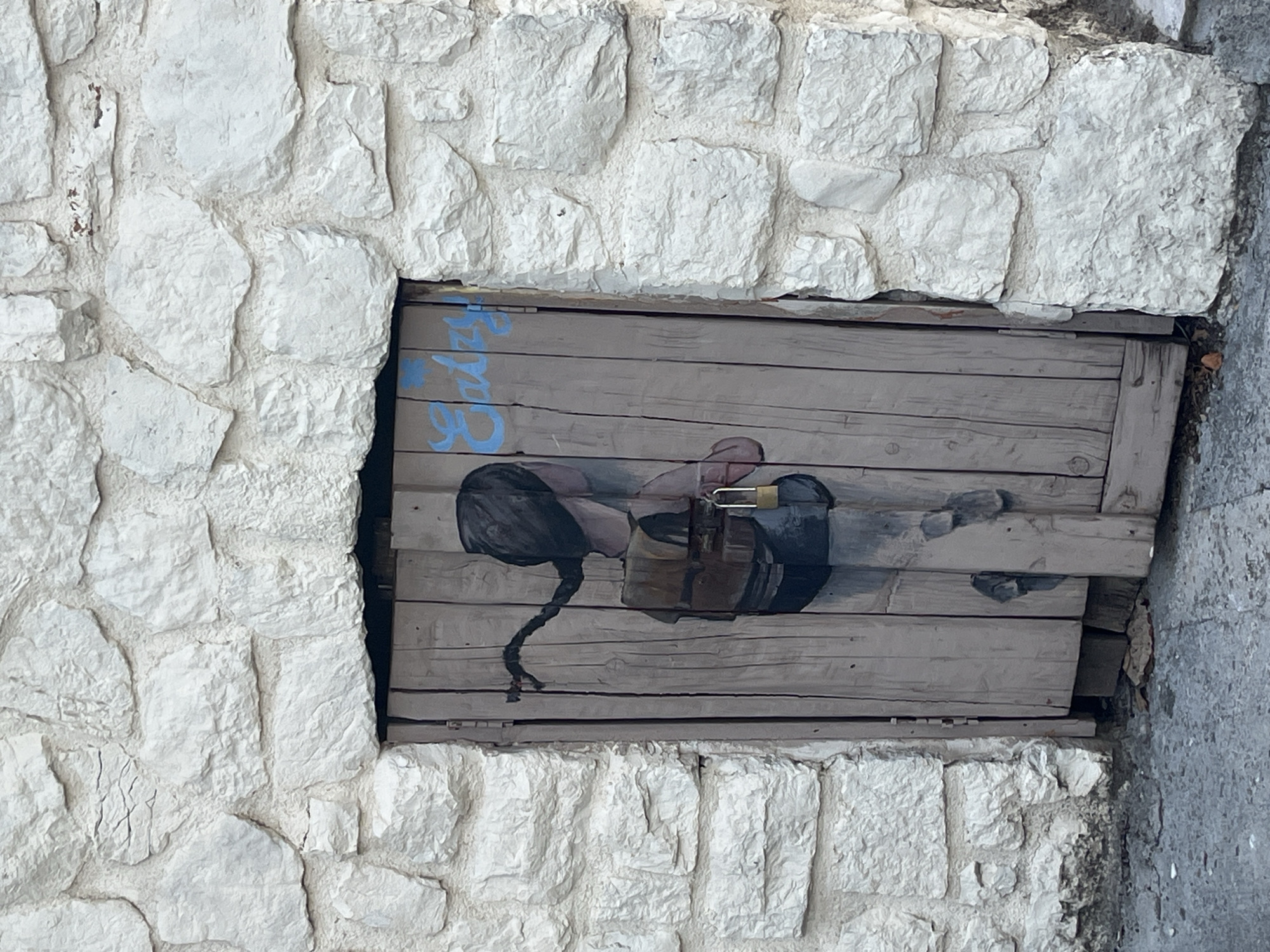 The image shows a wooden door set into a rough-hewn stone wall in Budva, Montenegro. The door is painted with a mural of a young girl with a braid and a backpack, and it is secured with a padlock. The stone wall is made of large, irregular white stones with visible mortar. The ground in front of the door is paved with grey concrete. The overall impression is one of an aged, rustic setting with a touch of street art.