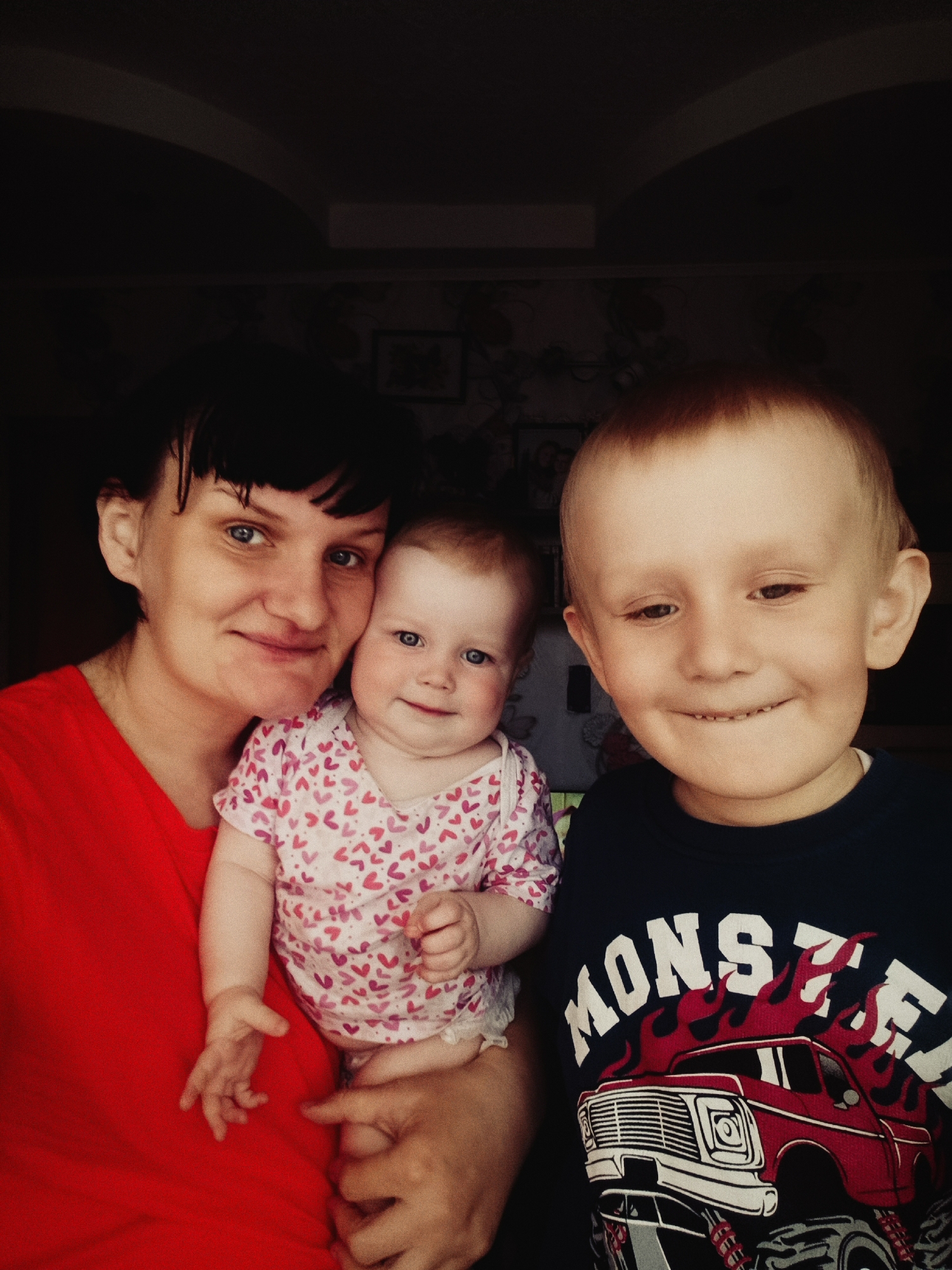 The image shows a woman, a baby, and a young child indoors. The woman, wearing a red shirt, is holding the baby, who is wearing a white onesie with pink and red hearts. A young boy is standing to the right, wearing a dark blue long-sleeve shirt with a monster truck design. The background appears to be a room with wallpaper or wall decorations. The location is the city of Unknown, Unknown.