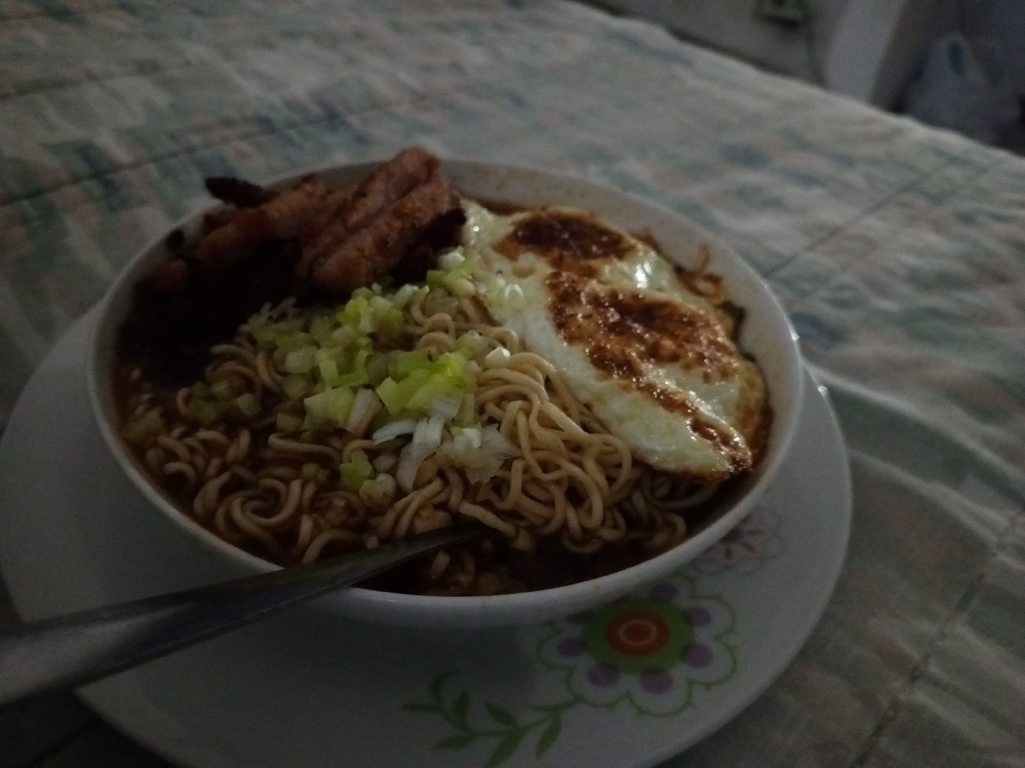 A bowl of ramen noodles sits on a patterned plate. The ramen is topped with chopped scallions, a fried egg with browned edges, and pieces of dark-colored meat. A spoon rests in the bowl. The plate is on a textured surface resembling a bedspread. The location is unspecified, and there is no evidence to suggest it is in Porlamar, Venezuela. There are no people visible.