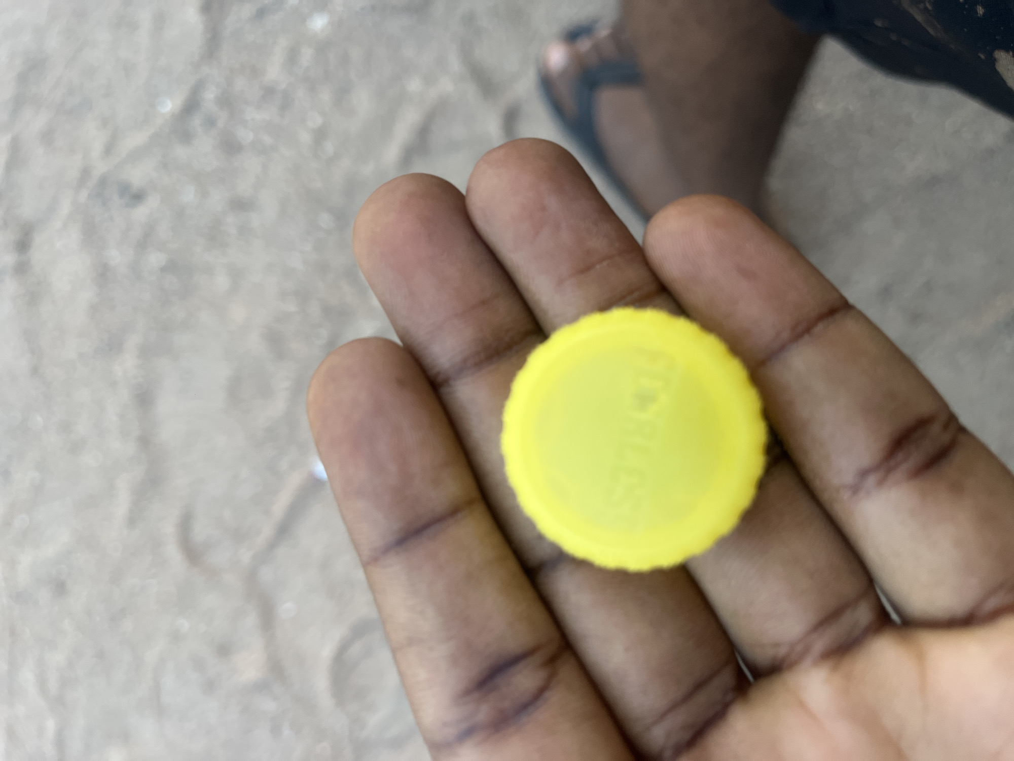 A close-up shot captures a yellow plastic bottle cap held in the palm of a person's hand. The cap has embossed text which appears to be a brand name. The hand is resting on a sandy surface, possibly a beach or a dry riverbed. In the background, out of focus, a person's leg wearing a sandal is visible, suggesting an outdoor setting in Jalingo, Nigeria. The lighting is natural and appears to be daylight.