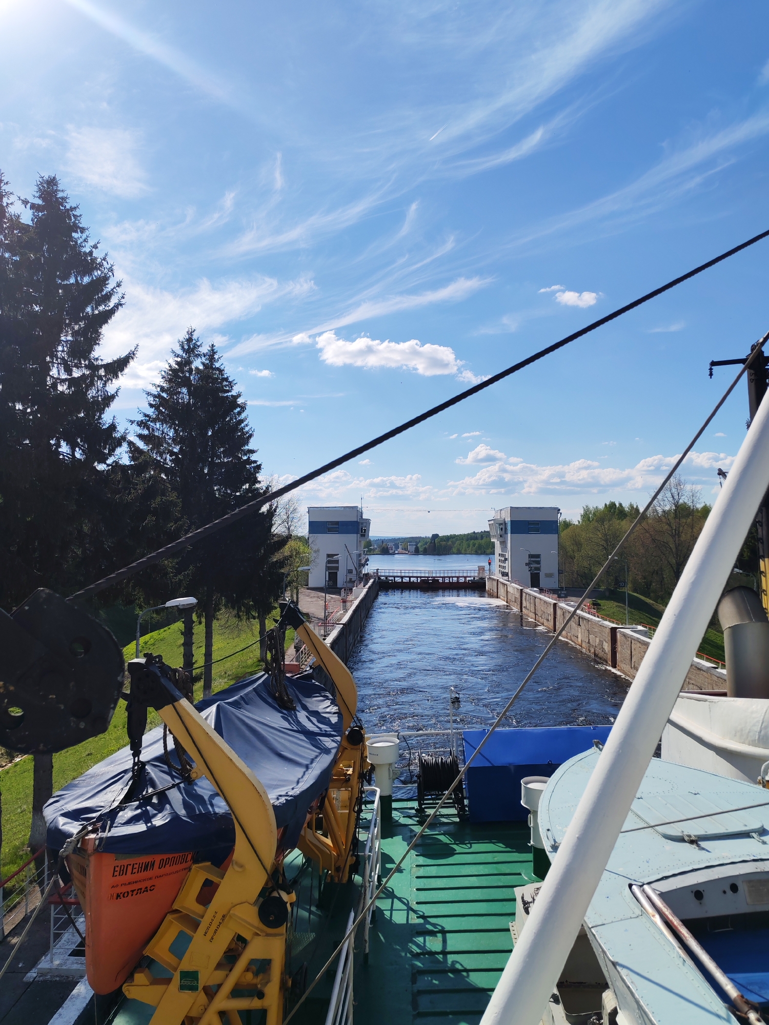 The image shows a view from a boat passing through a canal lock, likely in Alekseevskoe, Russia. The lock is defined by two white buildings on either side of the canal. In the foreground is a lifeboat on the boat. The sky is blue with some clouds. Trees can be seen along the banks of the canal. The water in the canal appears turbulent. A second boat is visible behind the first, within the lock.