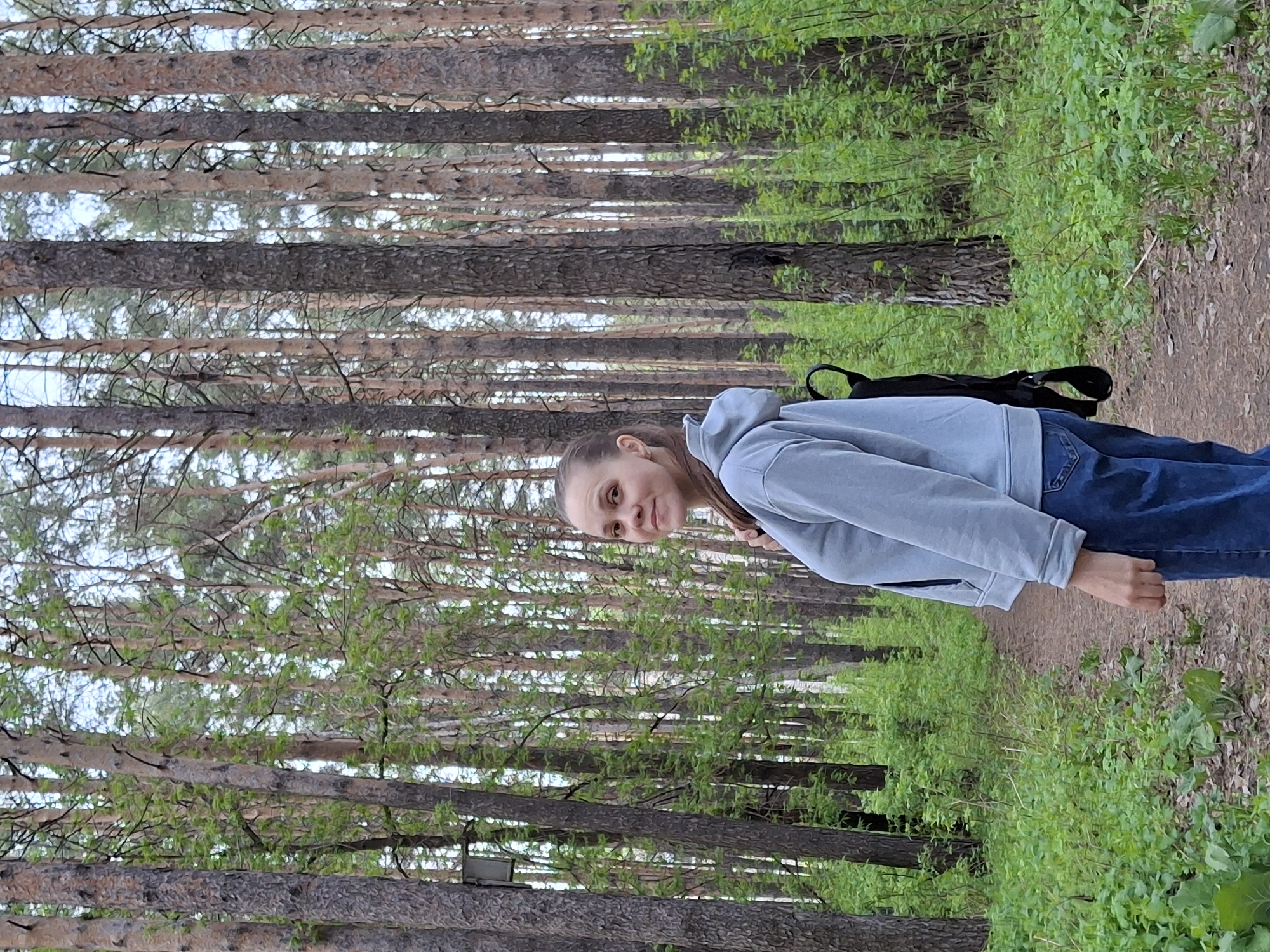A young woman stands on a dirt path in a forest. She wears a light blue hoodie, jeans, and a black backpack. The forest is dense with tall, thin trees. Green foliage grows along the path. The location is Chaikovsky, Russia.